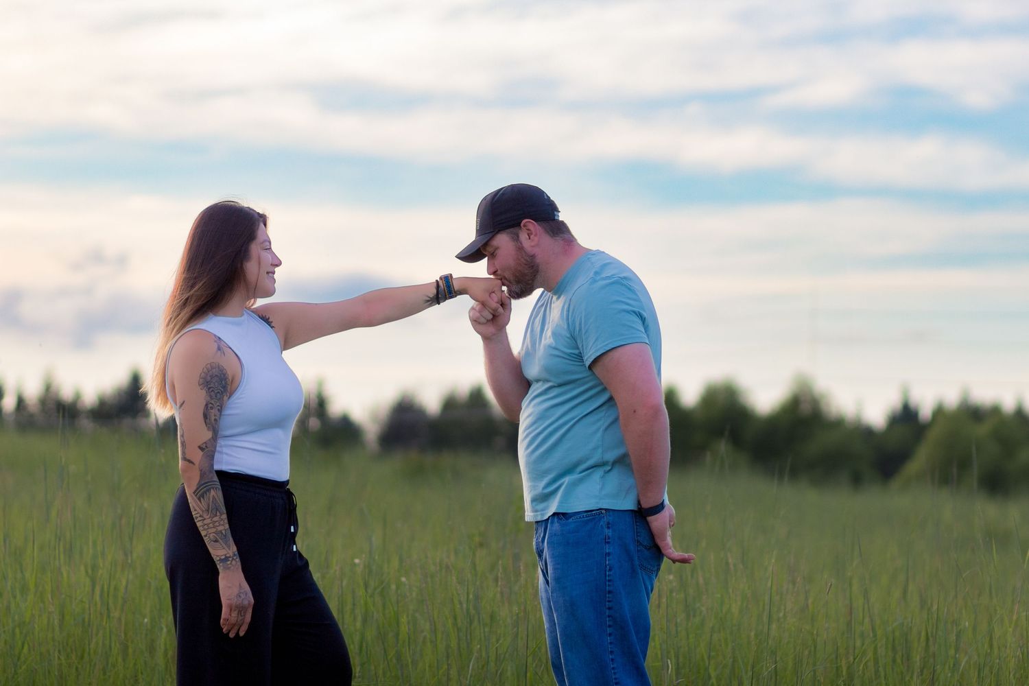 Two people share a tender moment in a green meadow with forest and cloudy sky as backdrop.