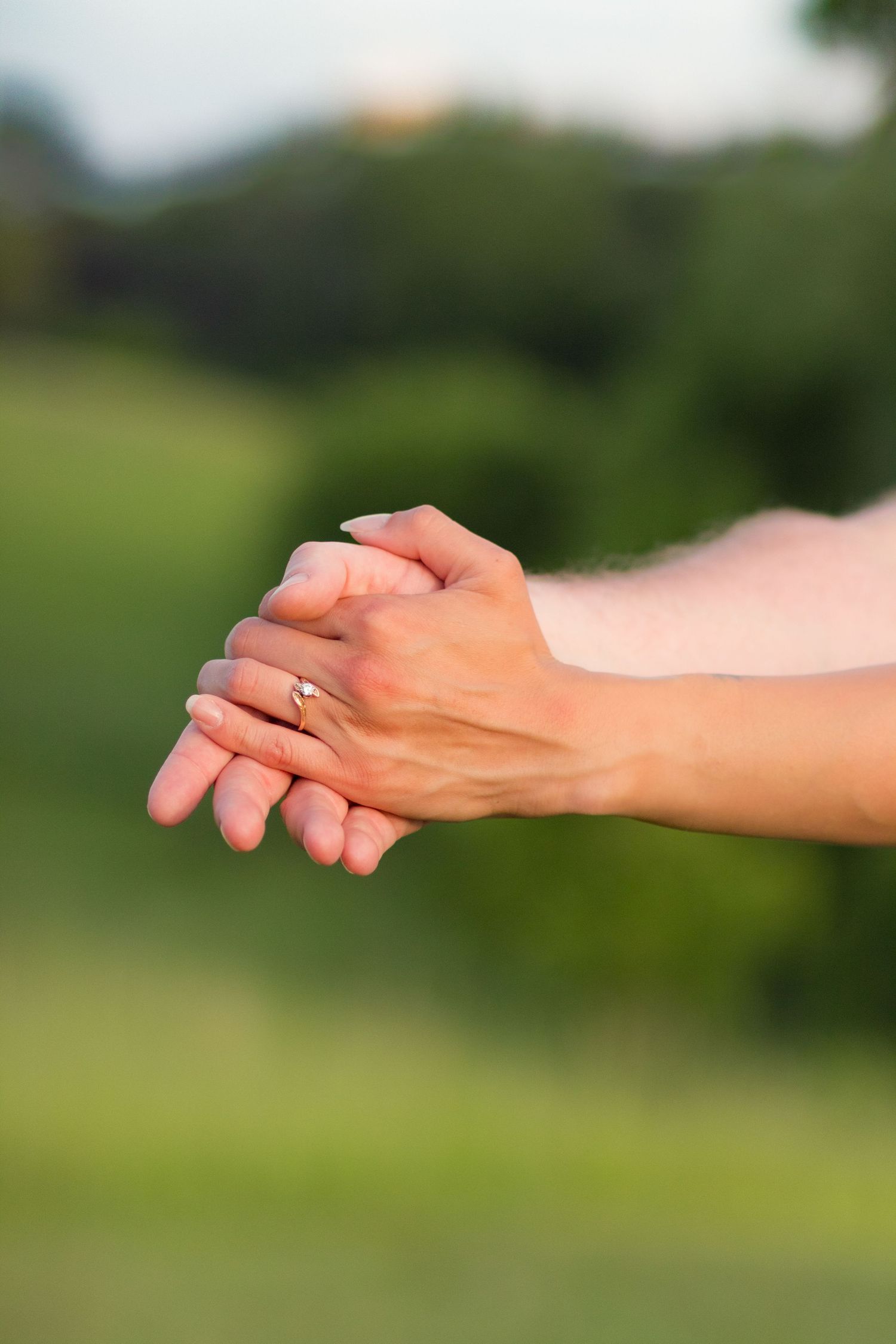 Close up of hands clasped together showing an engagement ring, with blurred green nature background outdoors.