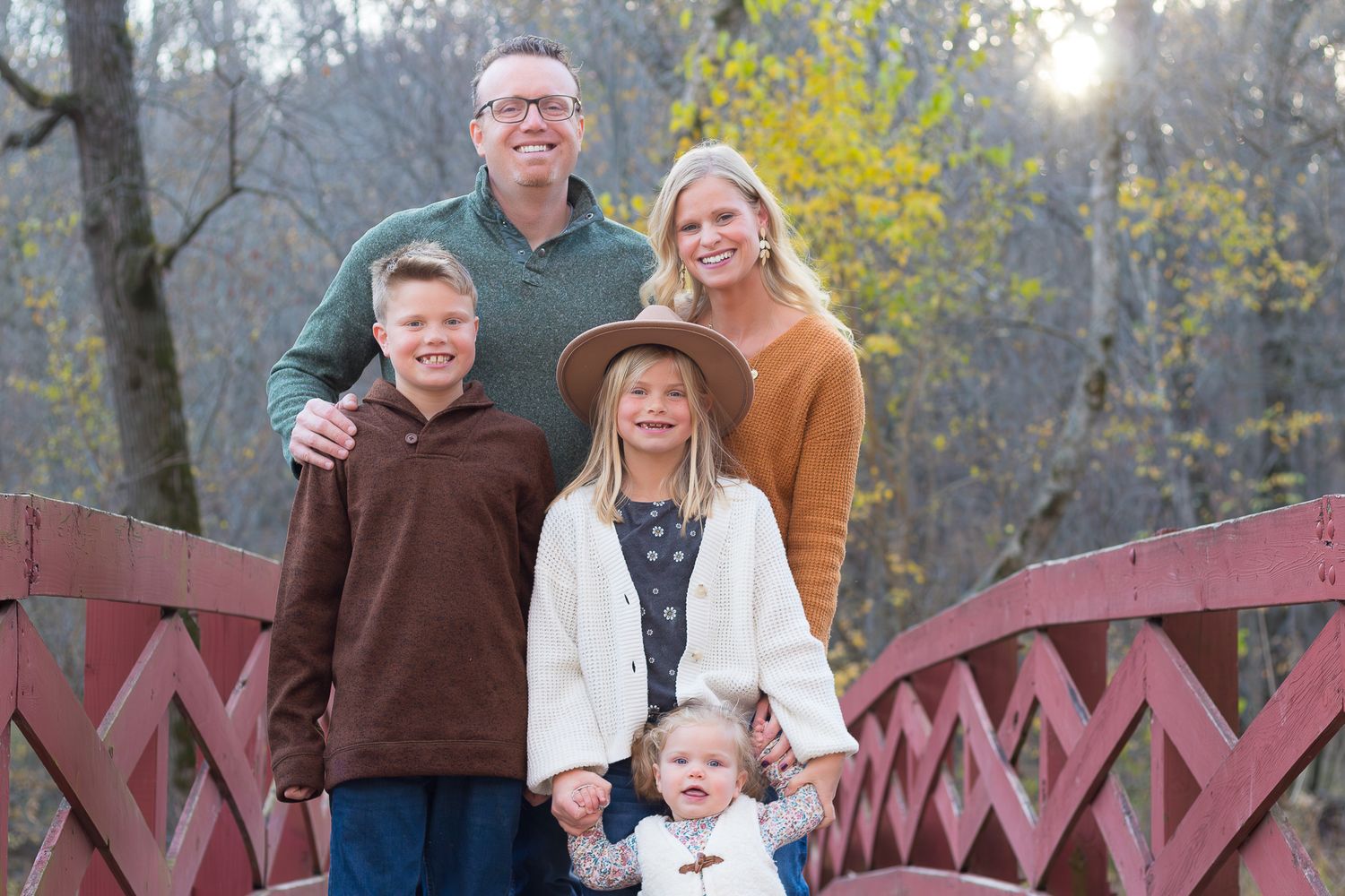 A family poses together on a red wooden bridge in an autumn forest setting.