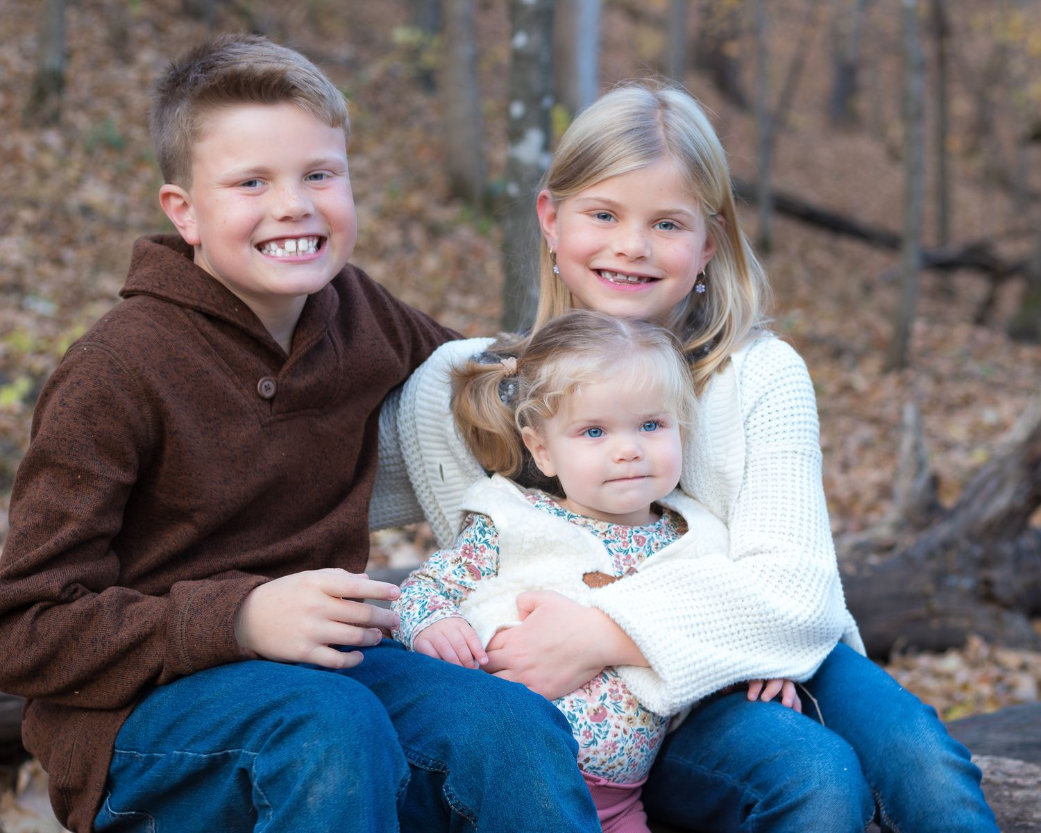 Three siblings sit close together for a portrait against a backdrop of fall foliage.
