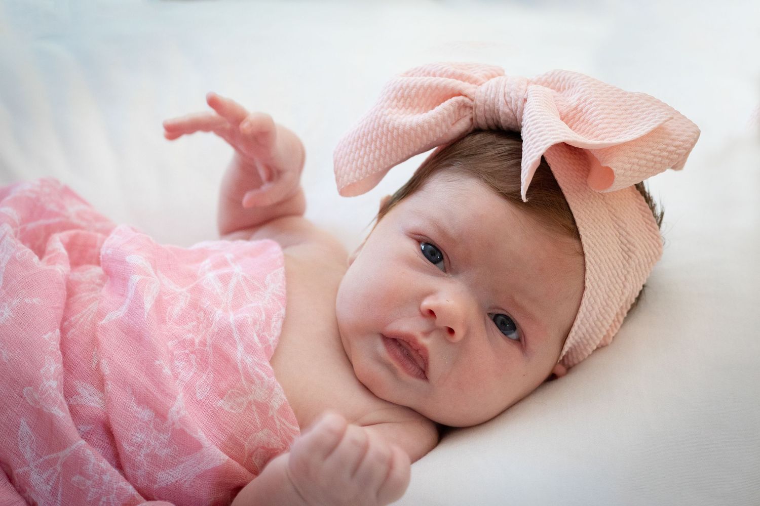Baby wearing pink outfit and bow headband lying on white bedding.