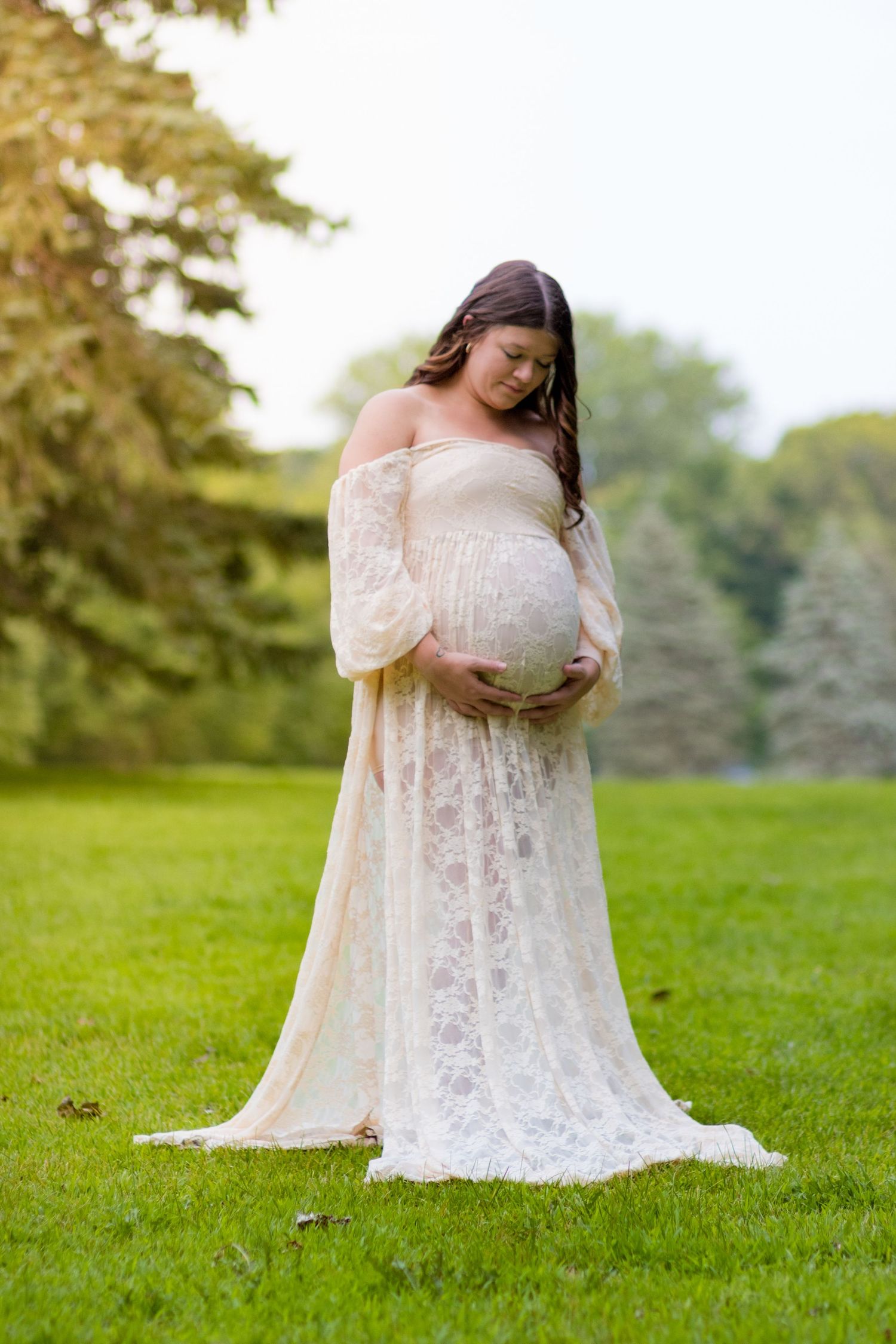 A pregnant woman in a flowing white lace maternity gown poses gracefully on a grassy lawn during an outdoor photoshoot.