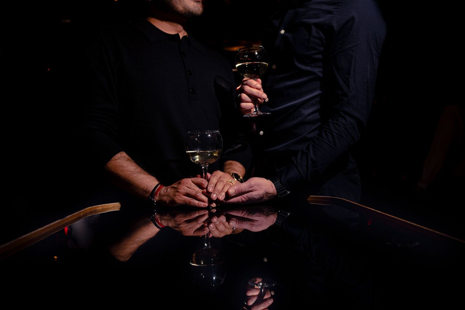 A dark moody photograph shows hands reflected on a glossy black surface with wine glasses in low light.