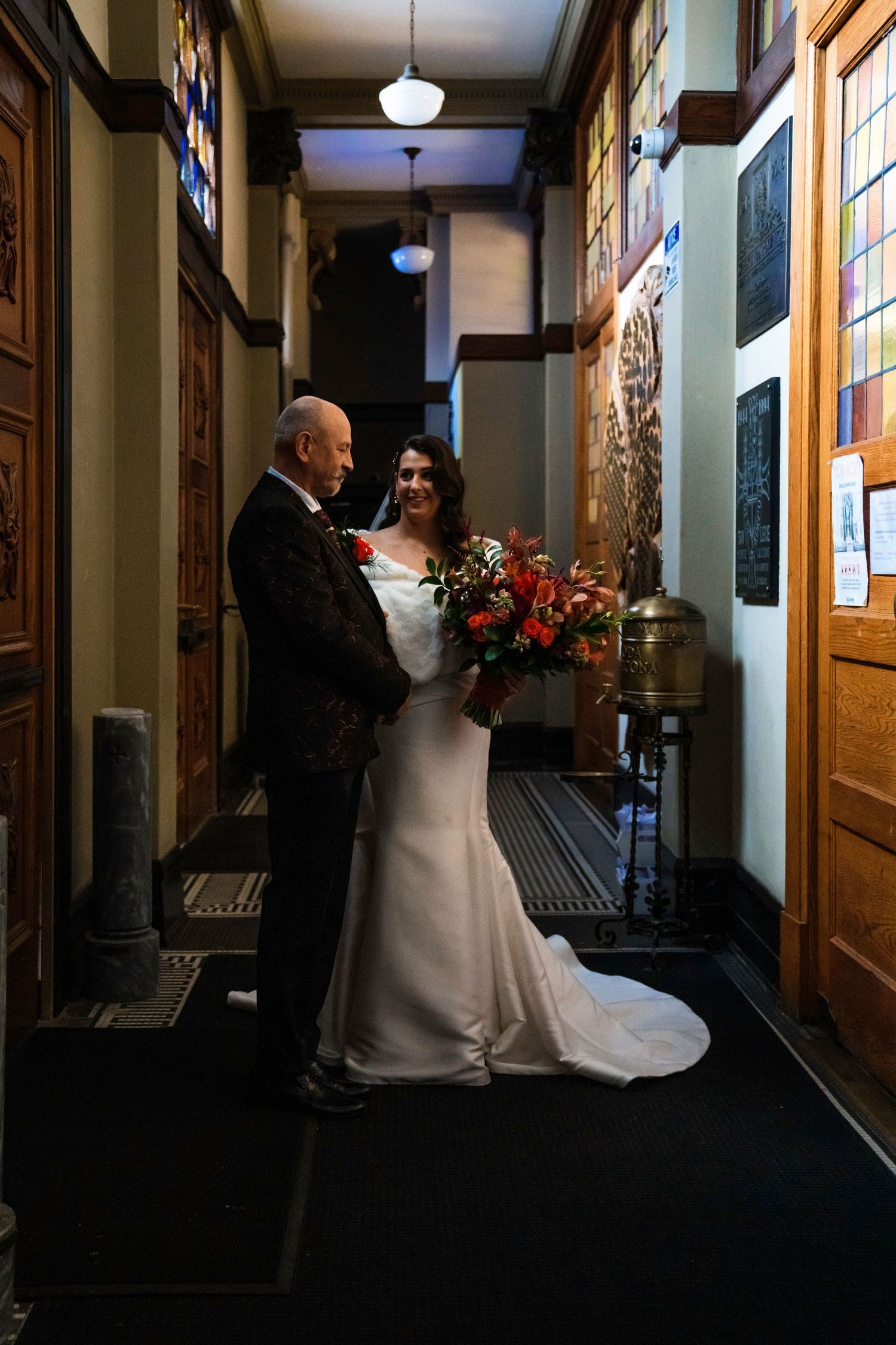 Intimate wedding portrait of bride and father of the bride in vintage hallway with warm lighting and floral bouquet.