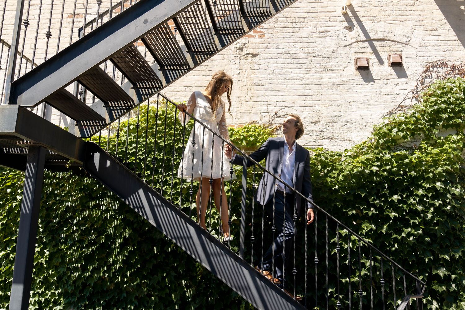 Couple exploring outdoor metal staircase with ivy-covered wall.