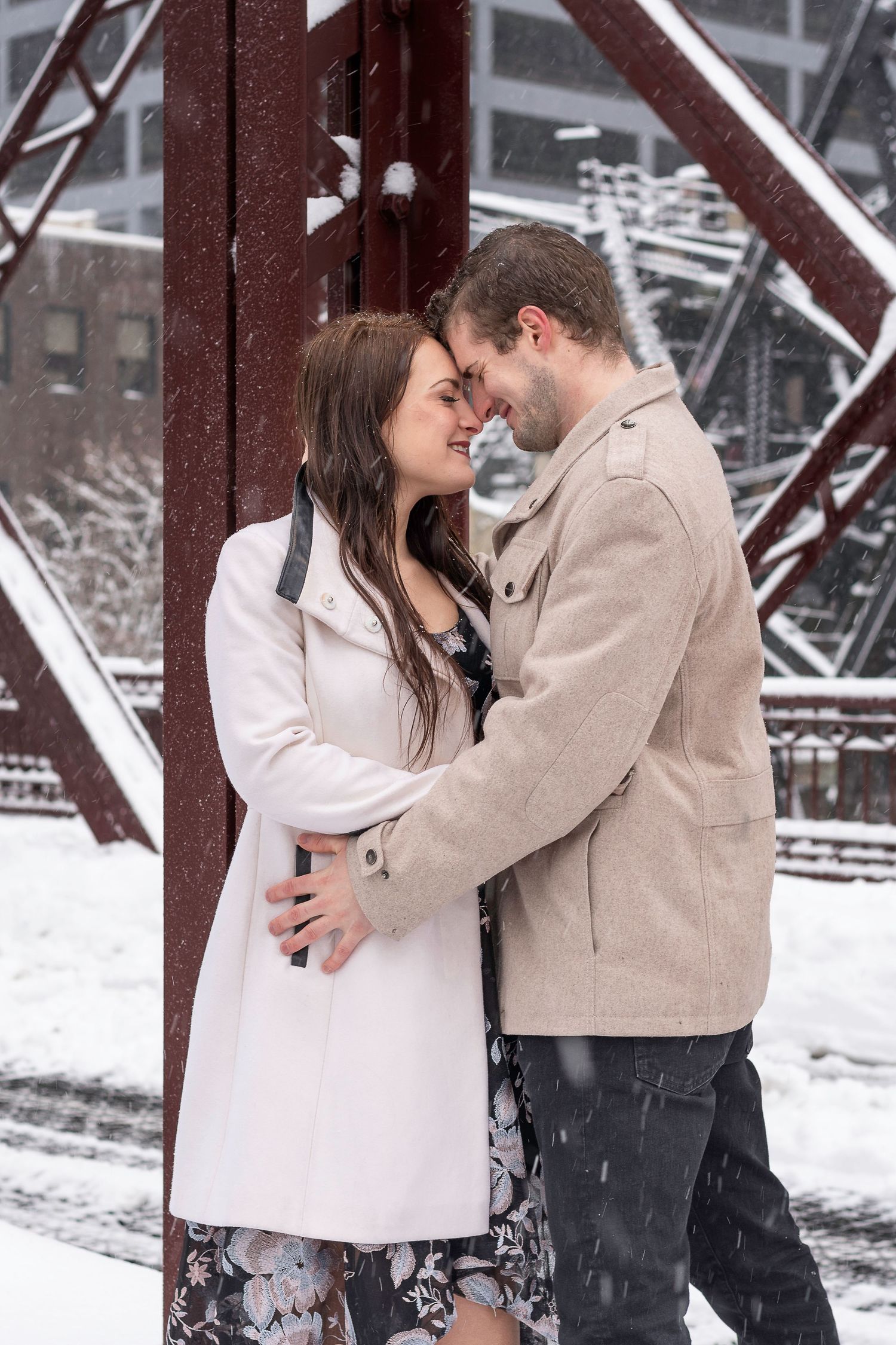 Romantic couple embracing in winter snow by red metal bridge structure.