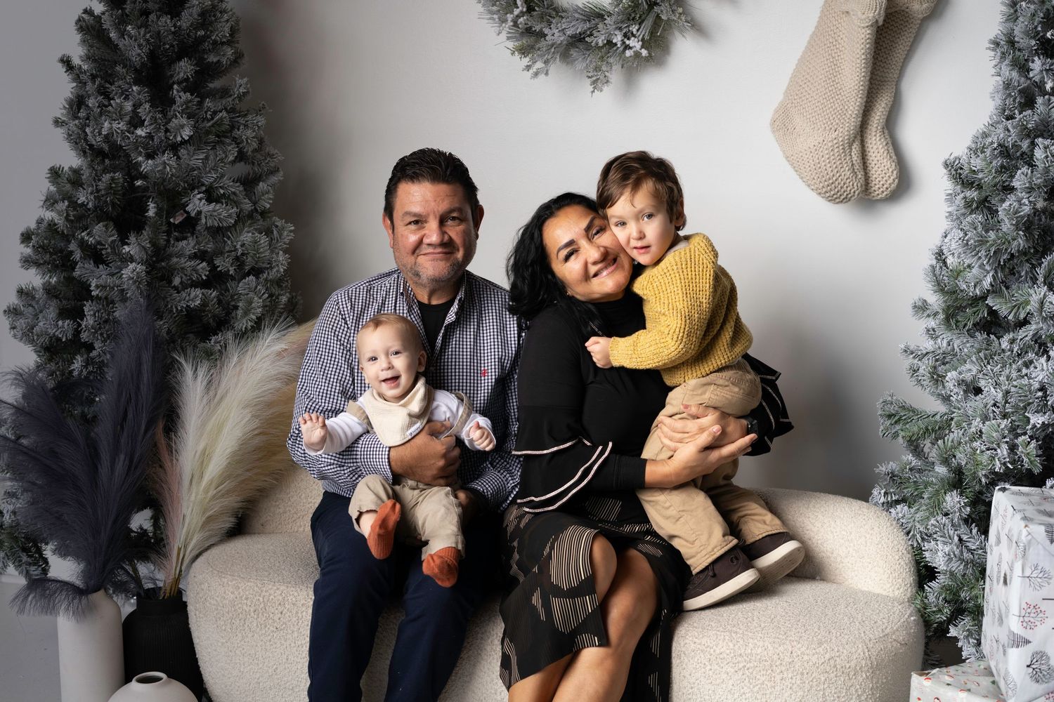 Family poses together for festive holiday photo with Christmas decor and stockings on wall.