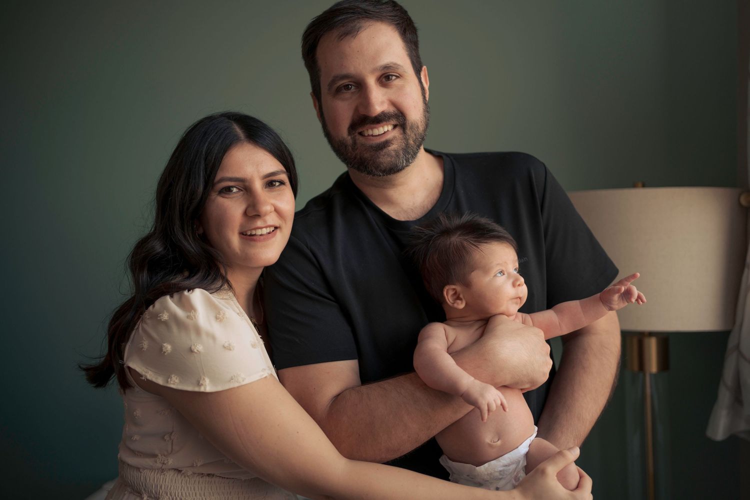 Family of three pose together in studio against teal wall.