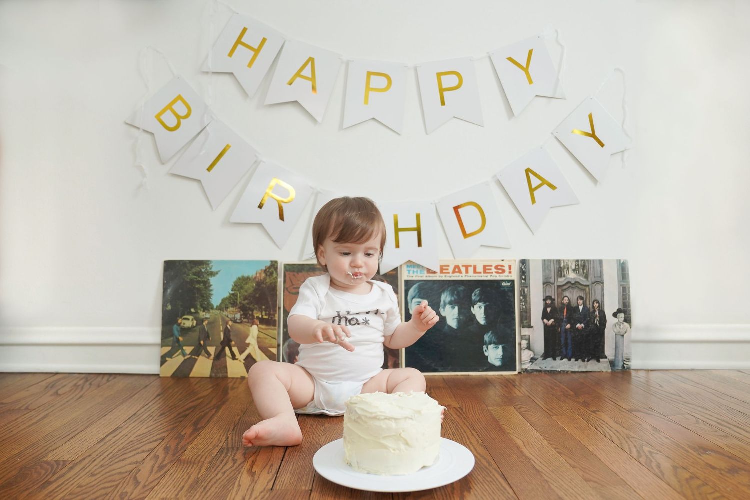 Baby in white onesie with birthday cake and Beatles albums display.