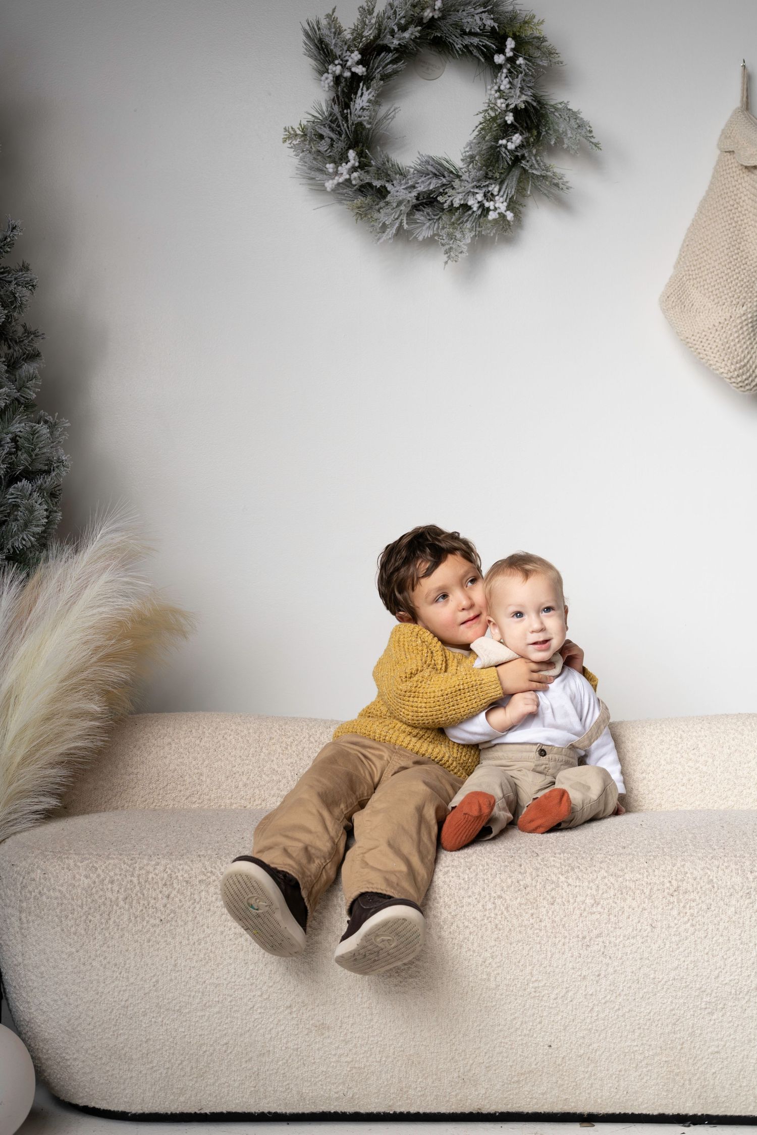 Two children sit together on beige couch with holiday wreath and Christmas tree decorations.