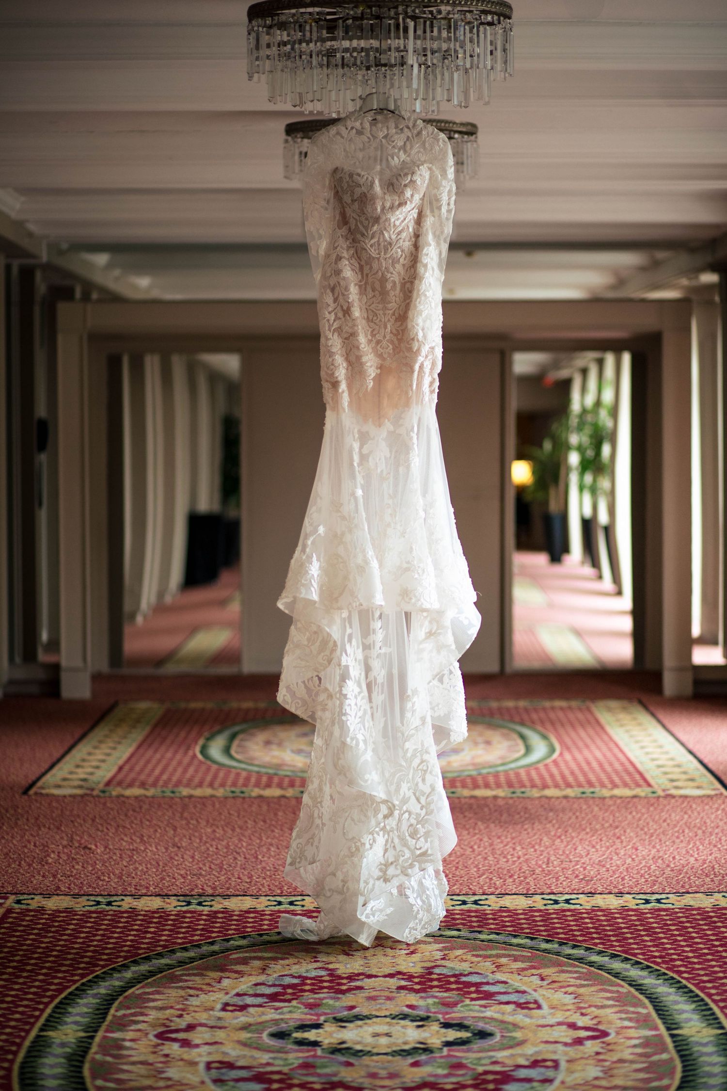 Elegant lace wedding dress hanging from crystal chandelier in luxurious hotel hallway.