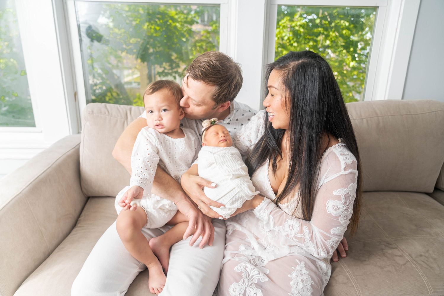 Family in white clothing sharing tender moment with baby on sofa by sunny window.