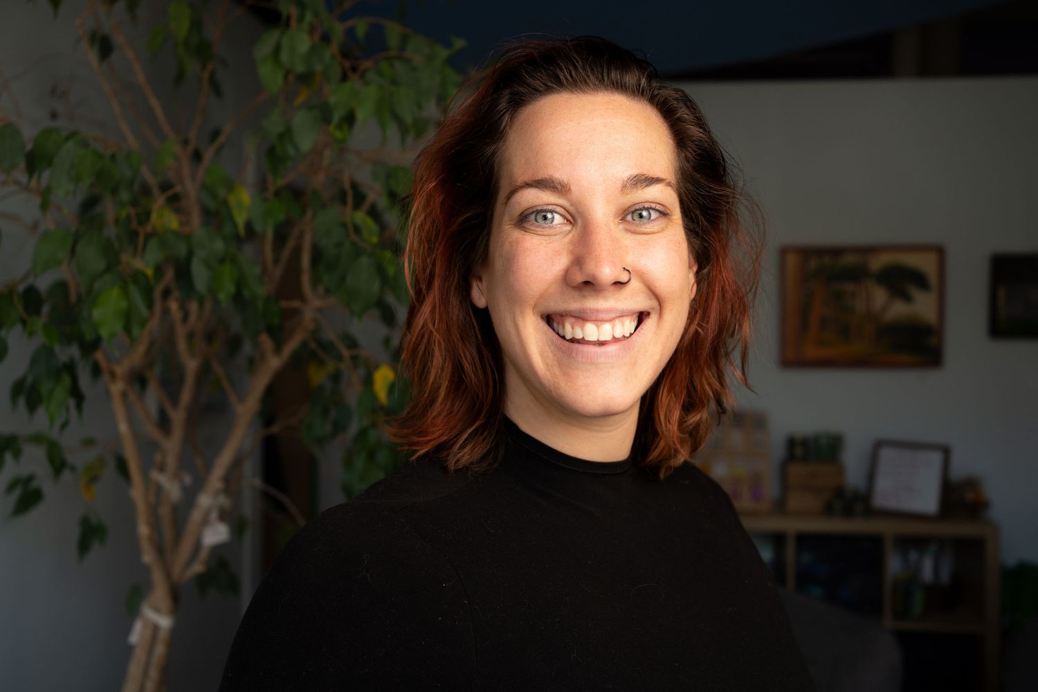 Person in black sweater smiling brightly against blurred interior background with green plant.  Headshot for yoga branding.