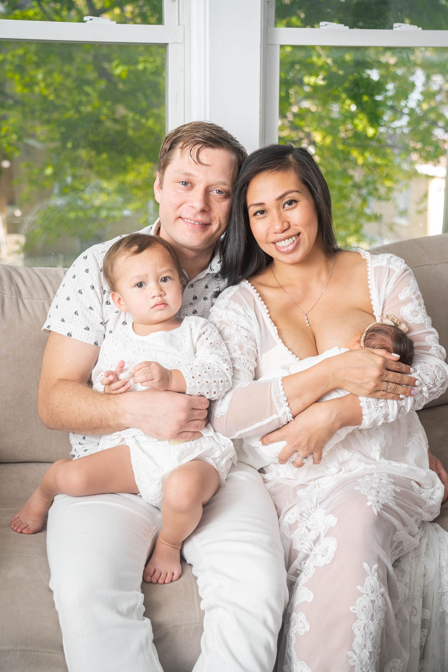 Family in white outfits sitting together on grey couch near bright window.
