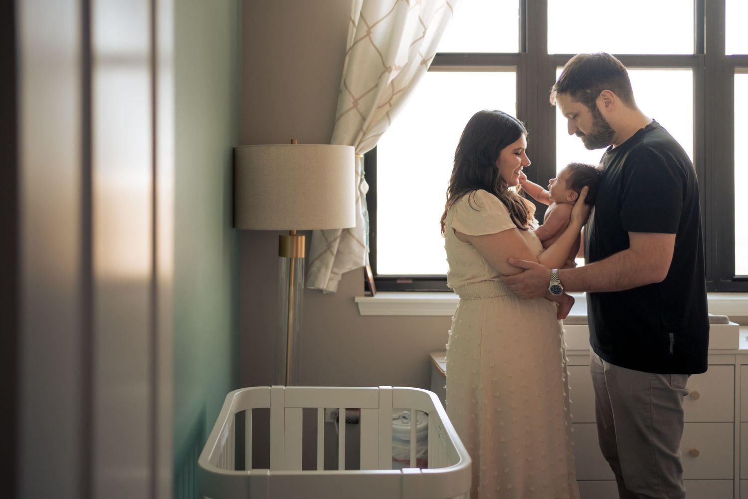 Parents holding newborn baby together in nursery with crib and lamp.