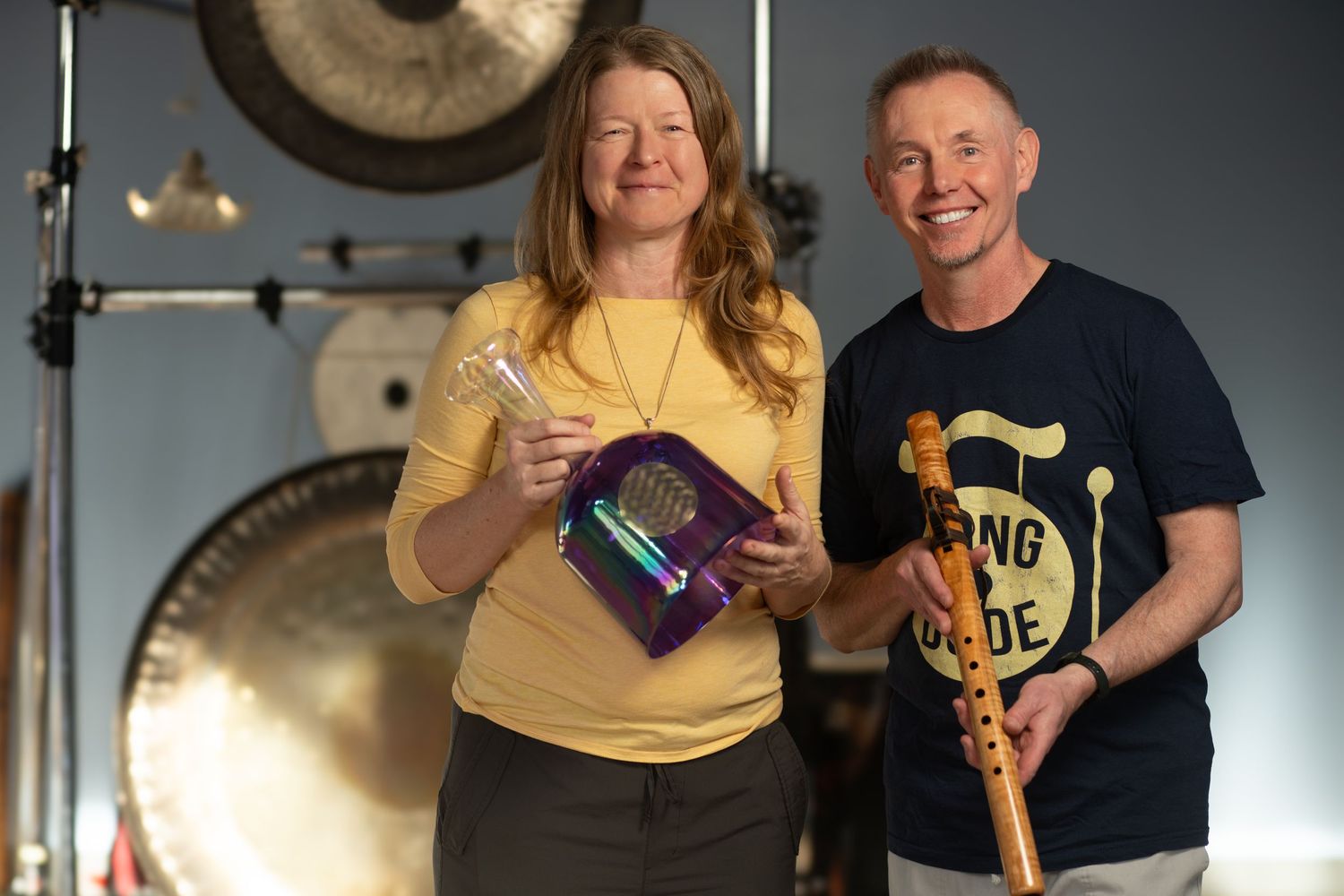 Two sound healers smiling and holding instruments in front of meditation gongs.