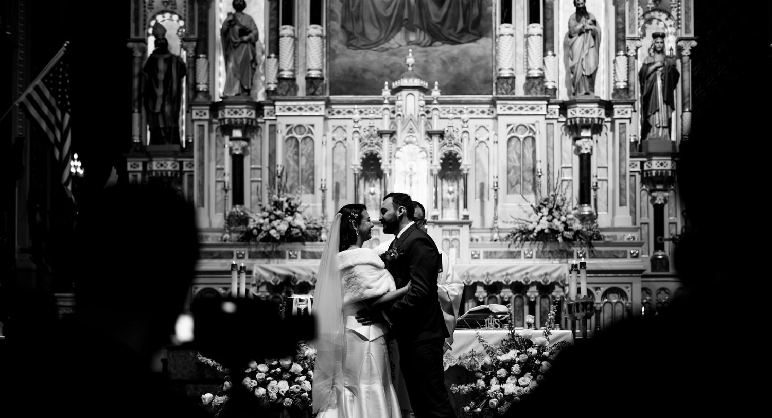 Black and white wedding ceremony photo in ornate church with dramatic altar backdrop.