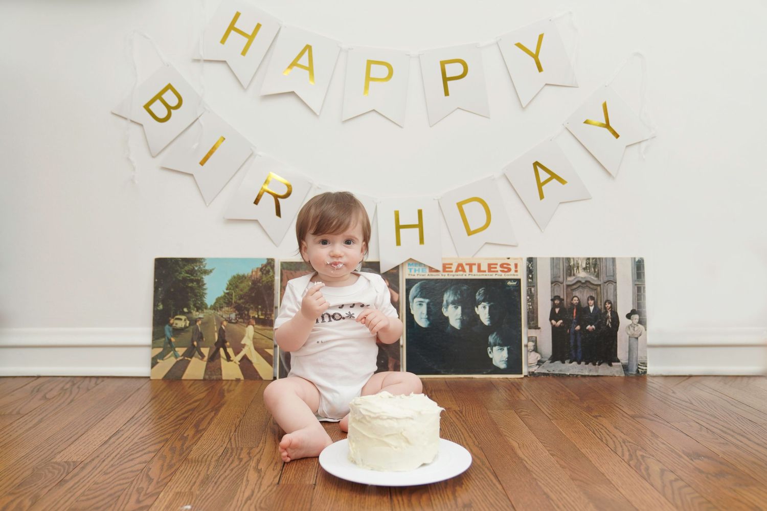 Baby sitting with birthday cake and Beatles vinyl records against wall with celebratory banner.