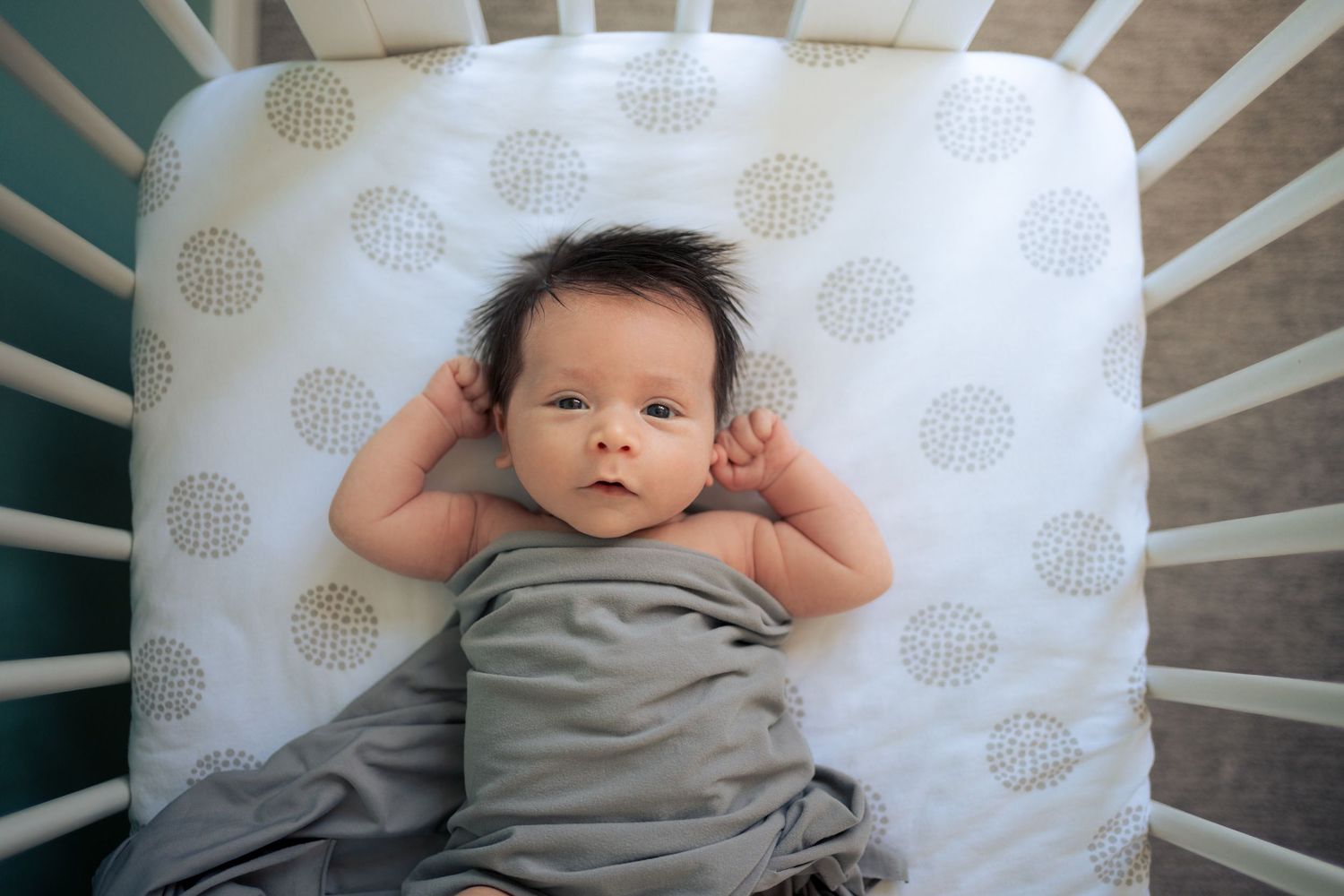 Baby lying in white polka dot crib wearing grey outfit.