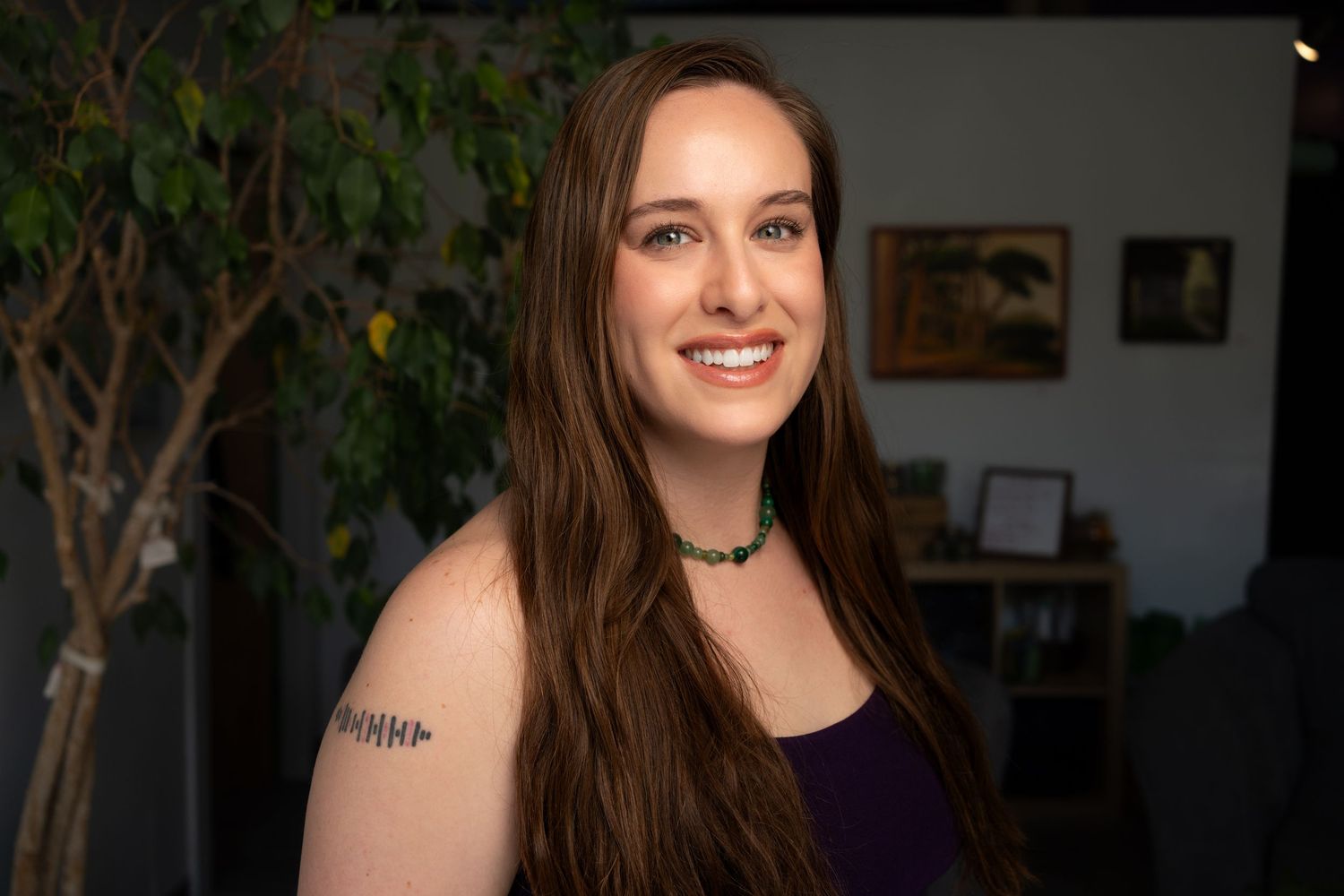 Portrait of woman with long brown hair wearing purple top standing in front of indoor plants. Headshot for yoga branding.