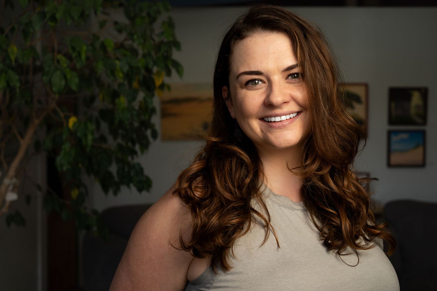 Person with flowing brown hair wearing grey top smiling warmly in indoor setting.  Headshot for yoga branding.