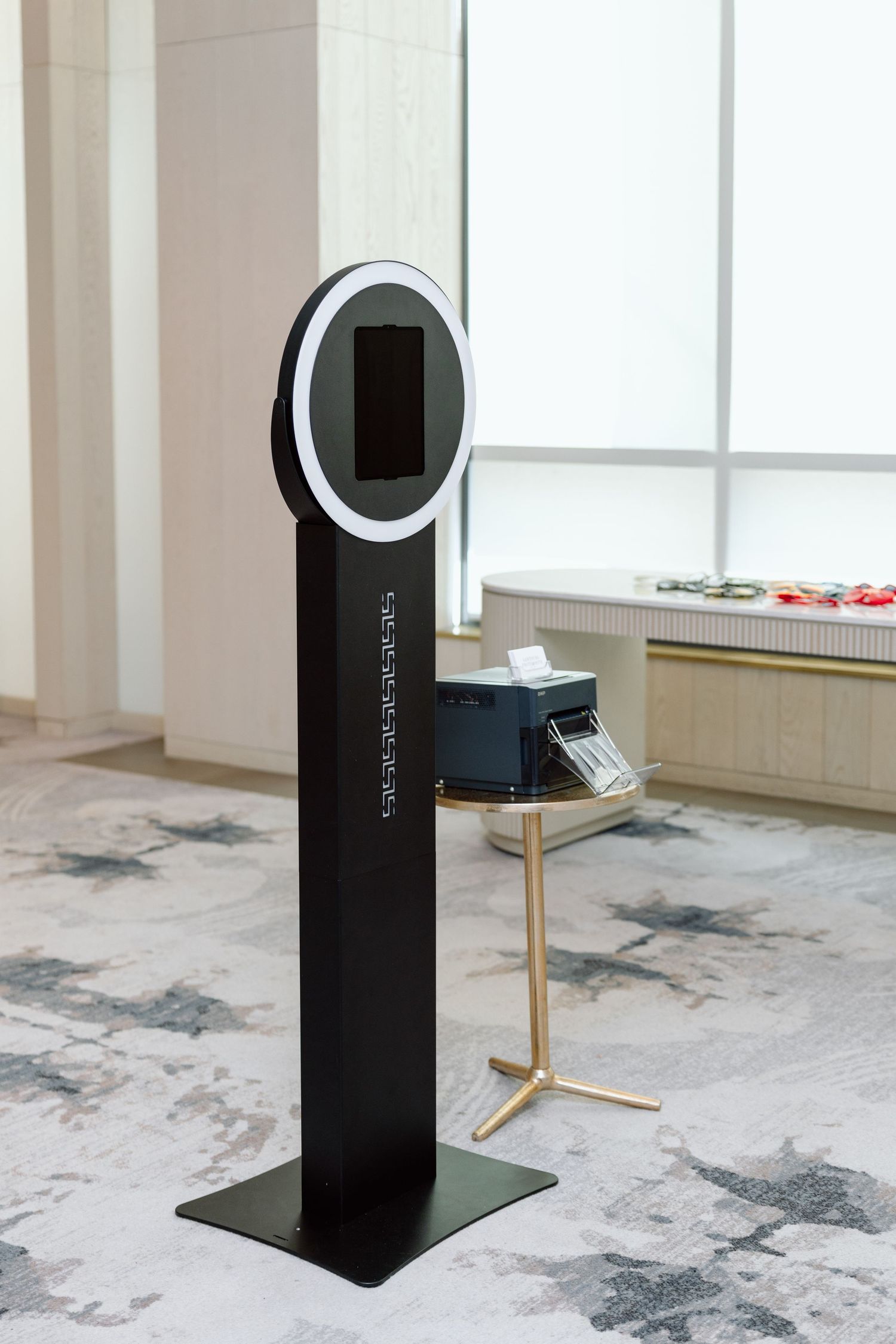 A sleek black photo booth kiosk with a circular ring light and touchscreen display stands on a patterned carpet in an elegant venue.