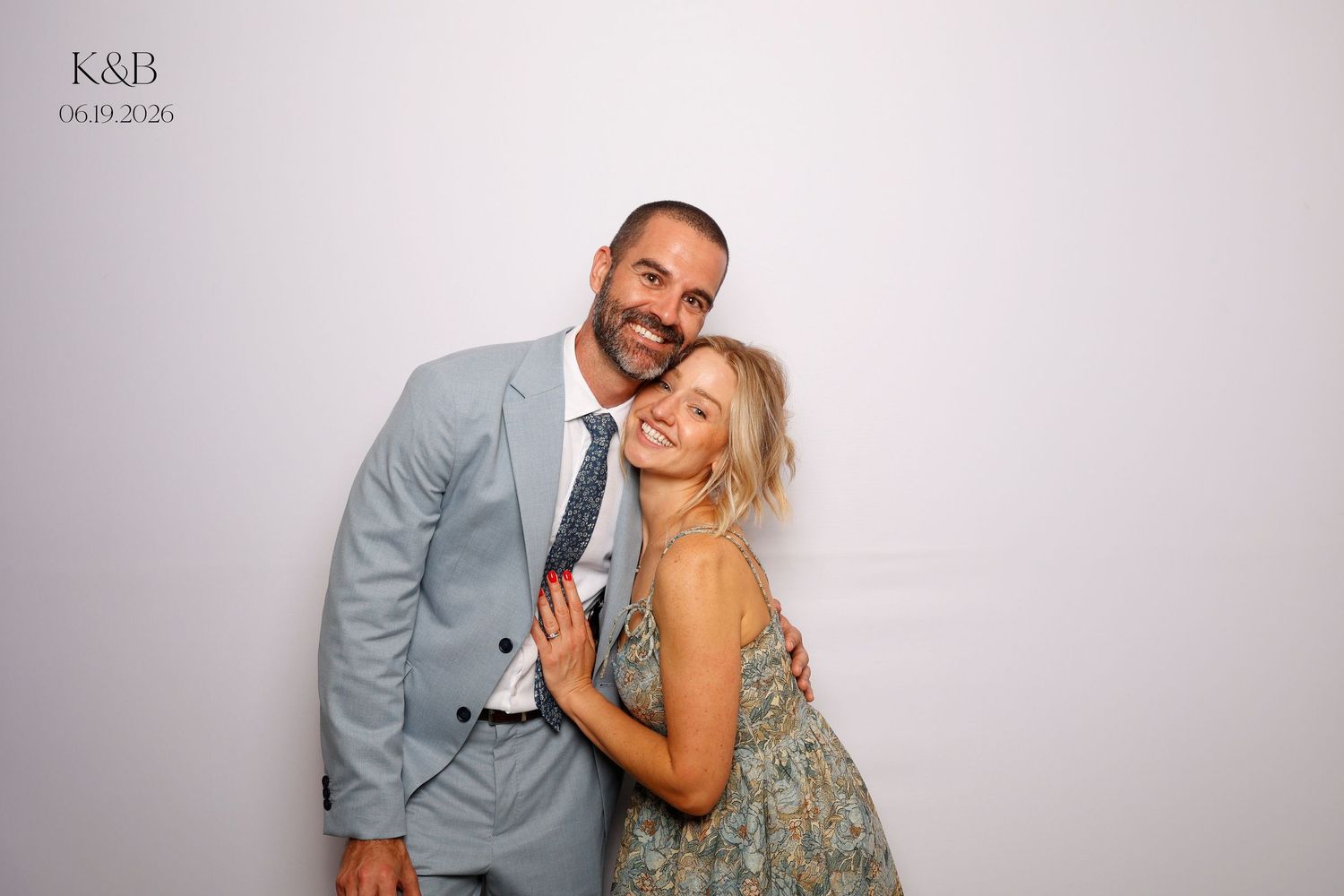 Couple posing together at a wedding photo booth, man in light gray suit, woman in floral dress, smiling warmly.