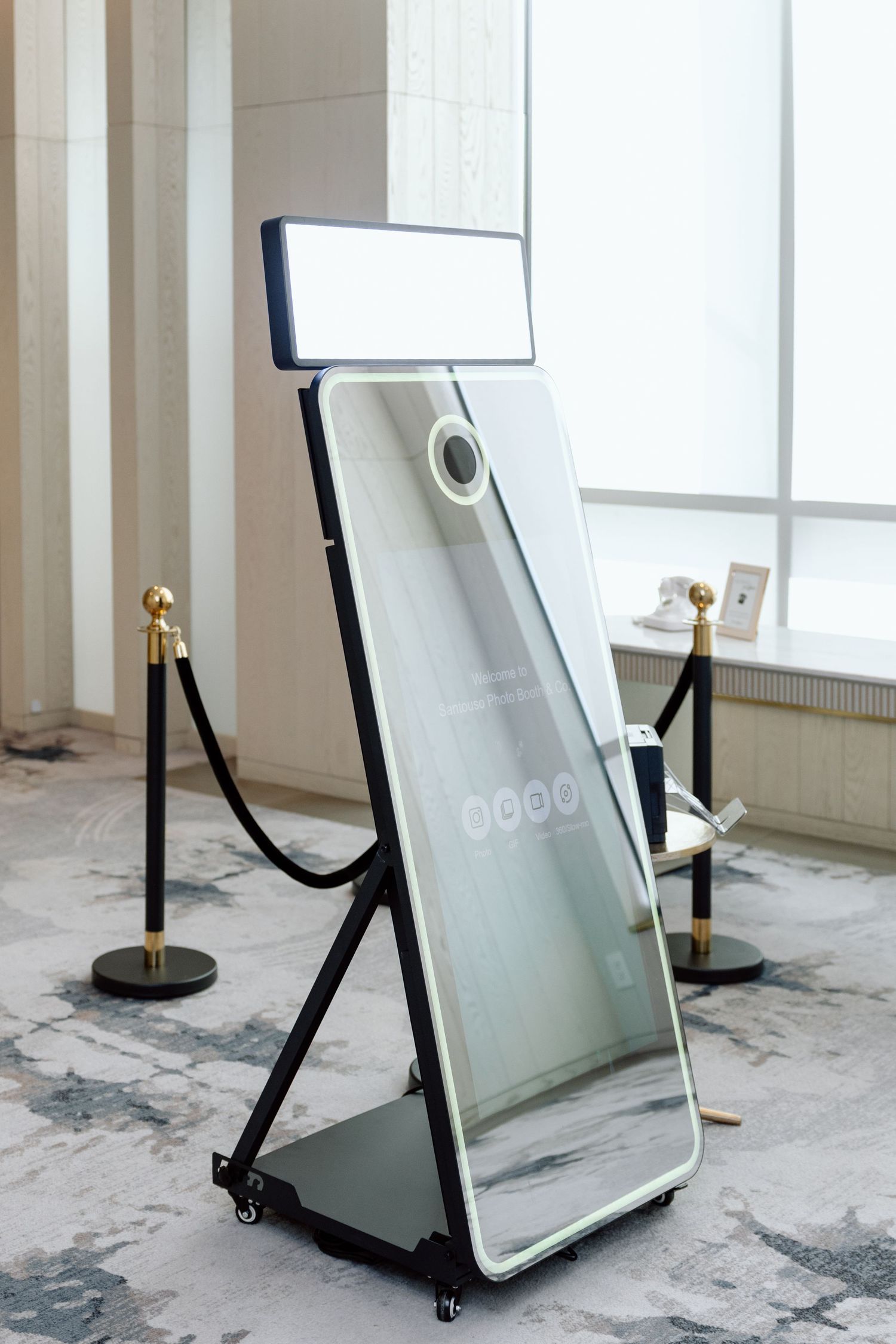 Modern mirror photo booth on wheels with LED light panel, framed by velvet rope stanchions in elegant venue.
