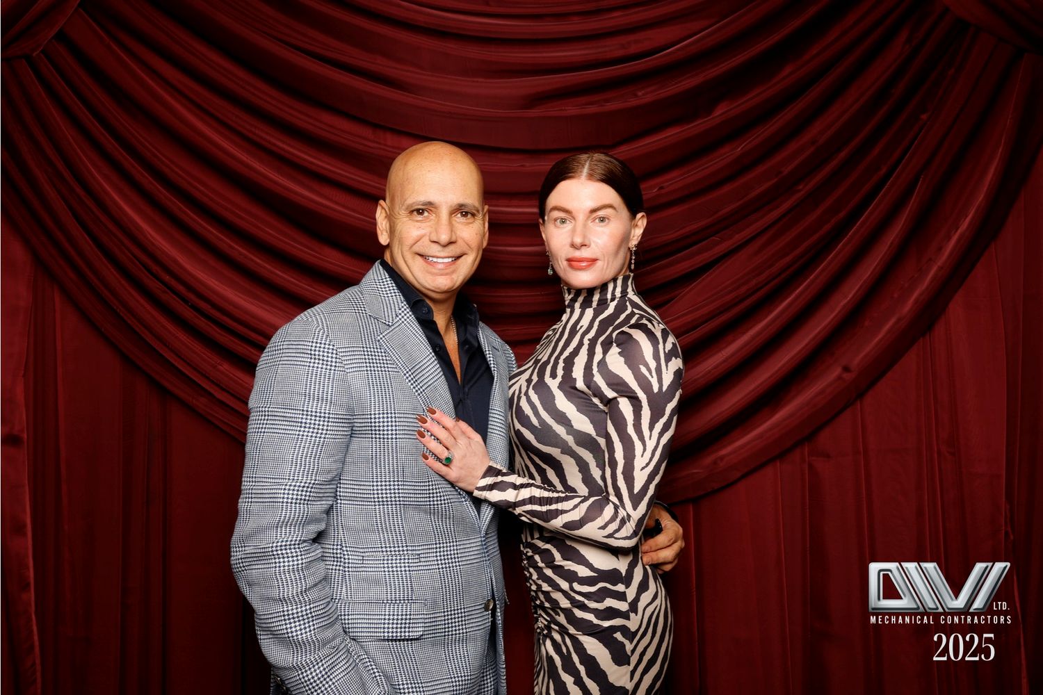 Two people pose together against a red curtain backdrop at a formal event.
