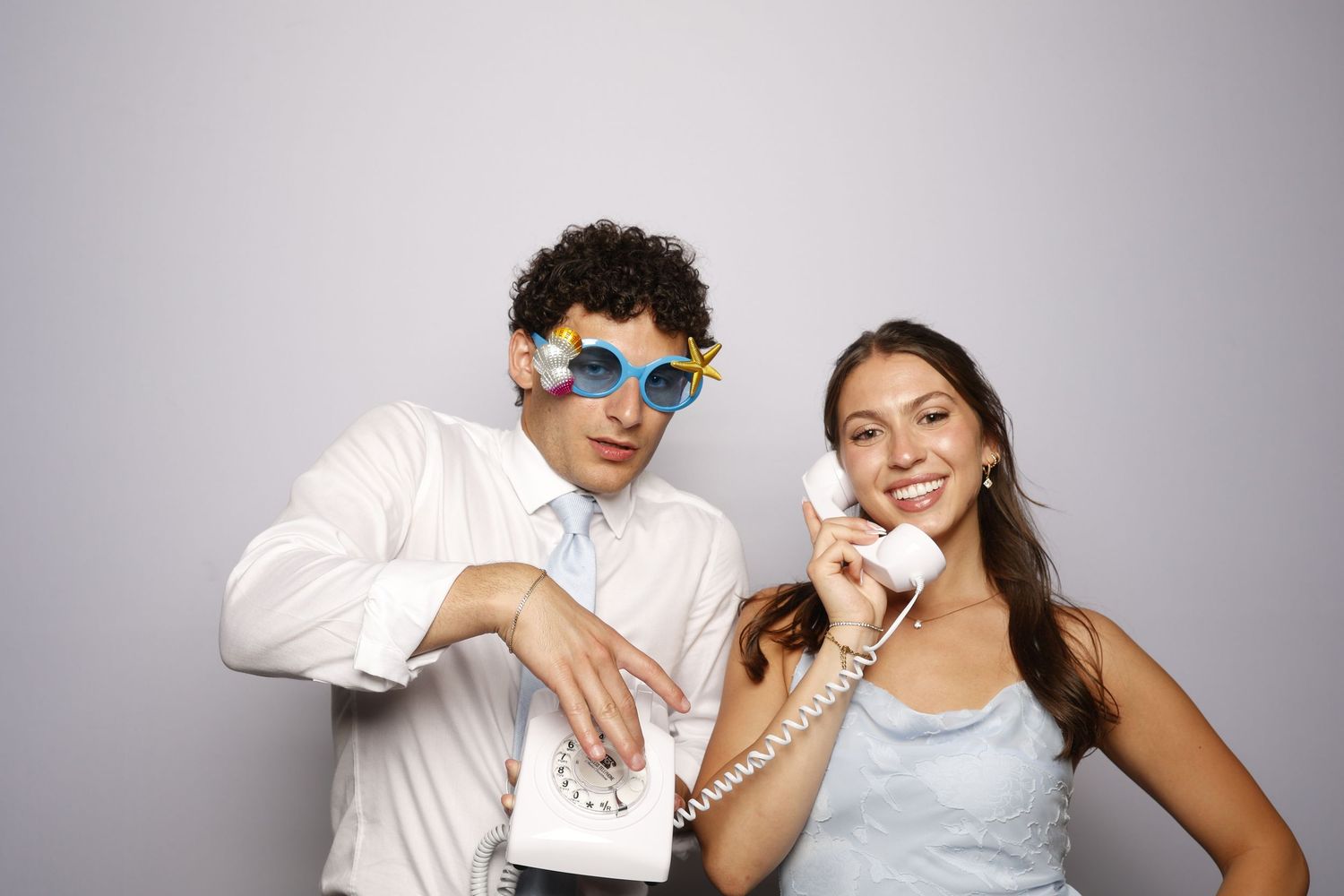 A young couple poses playfully in a photo booth, man wearing flower sunglasses holding a retro phone, woman smiling in blue dress.