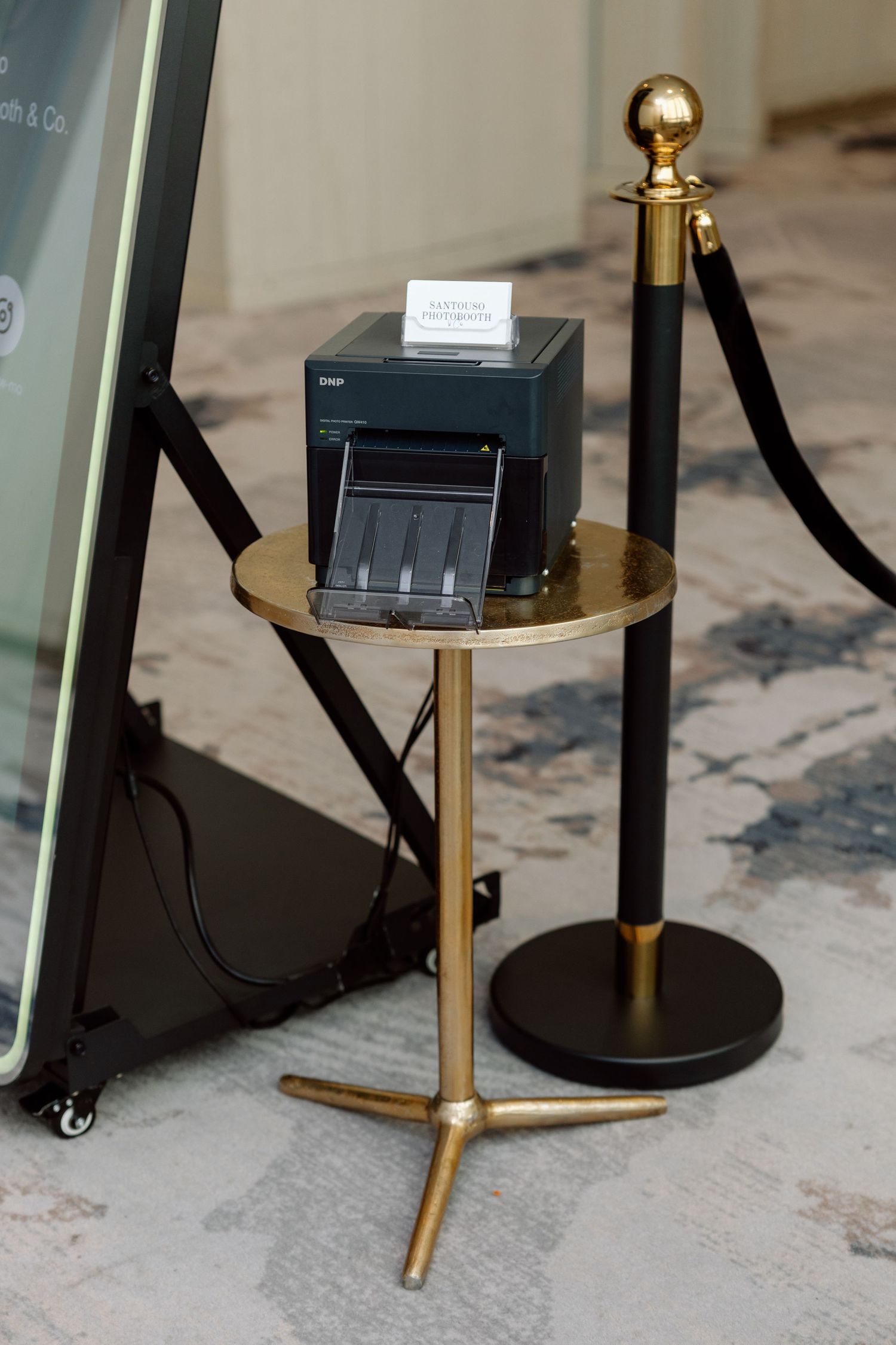 Gold pedestal table with a receipt printer behind velvet rope stanchions at an elegant event venue.