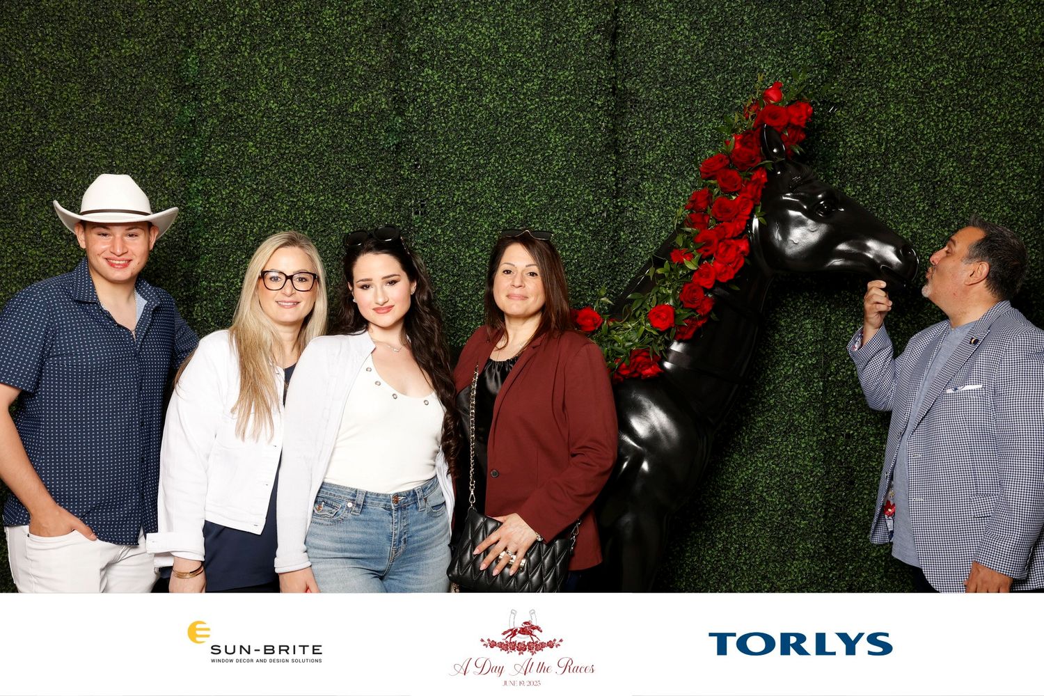 A group of people pose together against a green hedge backdrop at a sponsored event with company logos visible below.