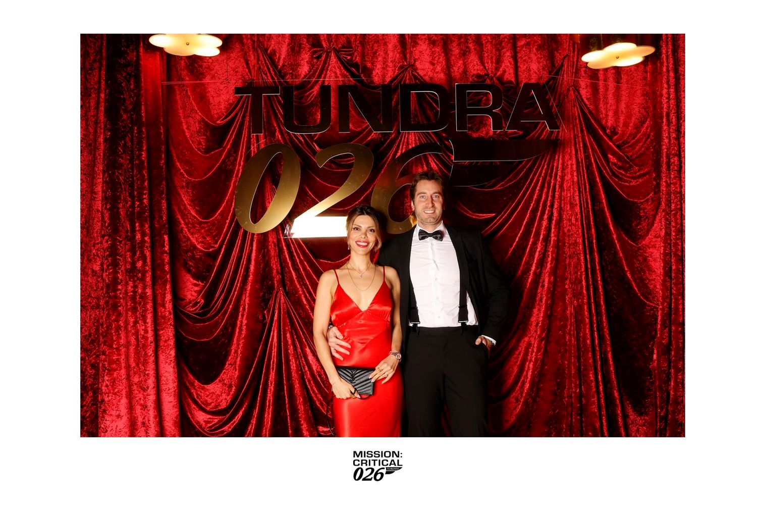 Figures in formal evening attire pose against a luxurious red velvet curtain backdrop at an elegant event.