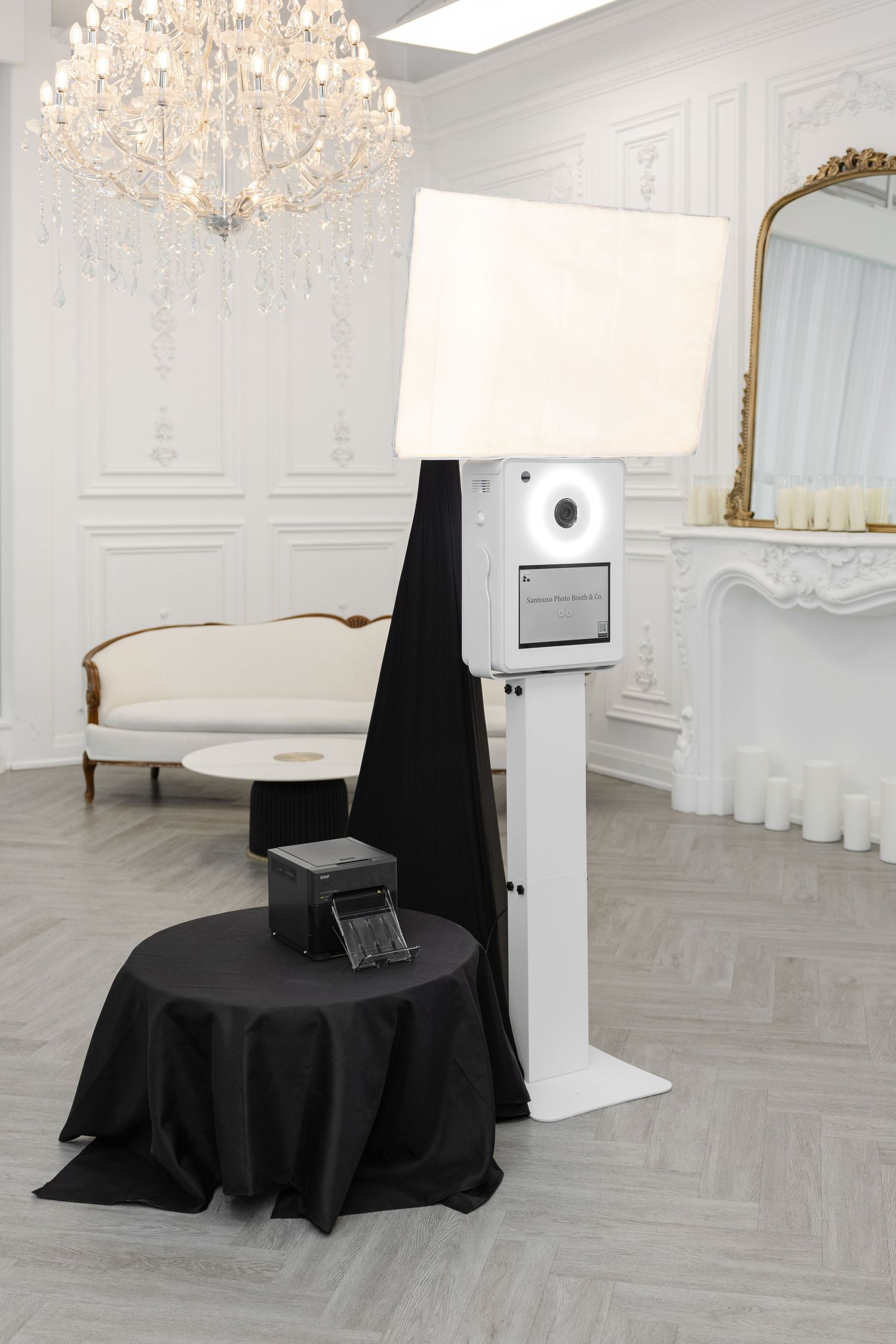 Elegant photo booth setup with crystal chandelier, white chaise lounge, and black draped table in a luxurious white room.