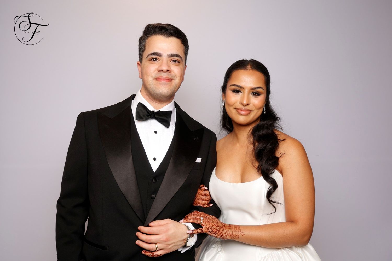 A couple in formal black tie attire poses together for a series of wedding portraits against a gray backdrop.