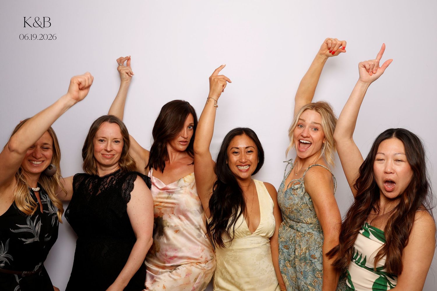 Six women in elegant dresses celebrate joyfully at a K&B wedding photo booth, arms raised and smiling on June 19, 2026.