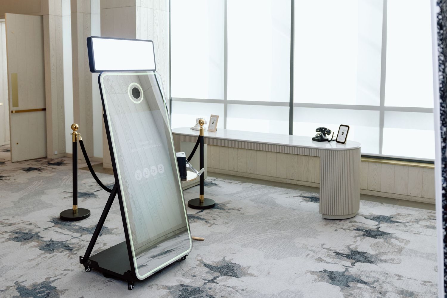 Modern mirror photo booth with LED light on elegant patterned carpet in bright event venue with large windows.