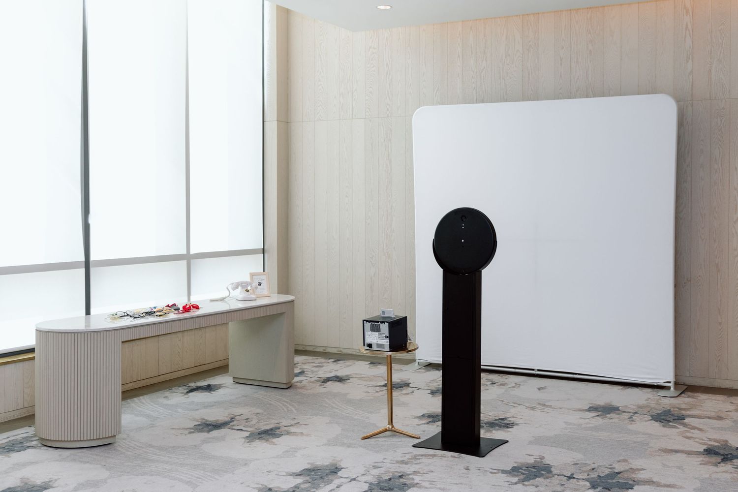 Modern minimalist room with white backdrop, sleek speaker on stand, and curved desk near large windows.