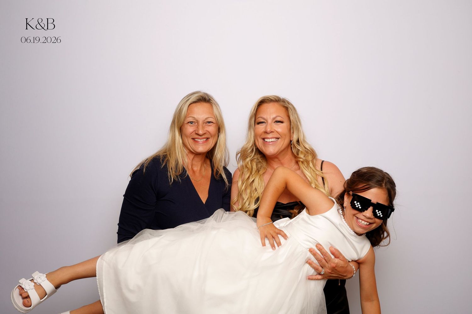 Three women pose playfully at a K&B wedding photo booth, with one woman being carried in a white dress.