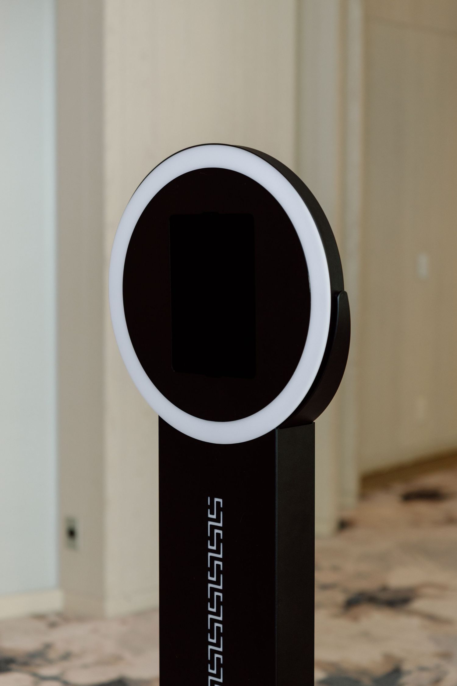 Modern black photo booth kiosk with circular ring light on a pedestal stand in an elegant hallway.