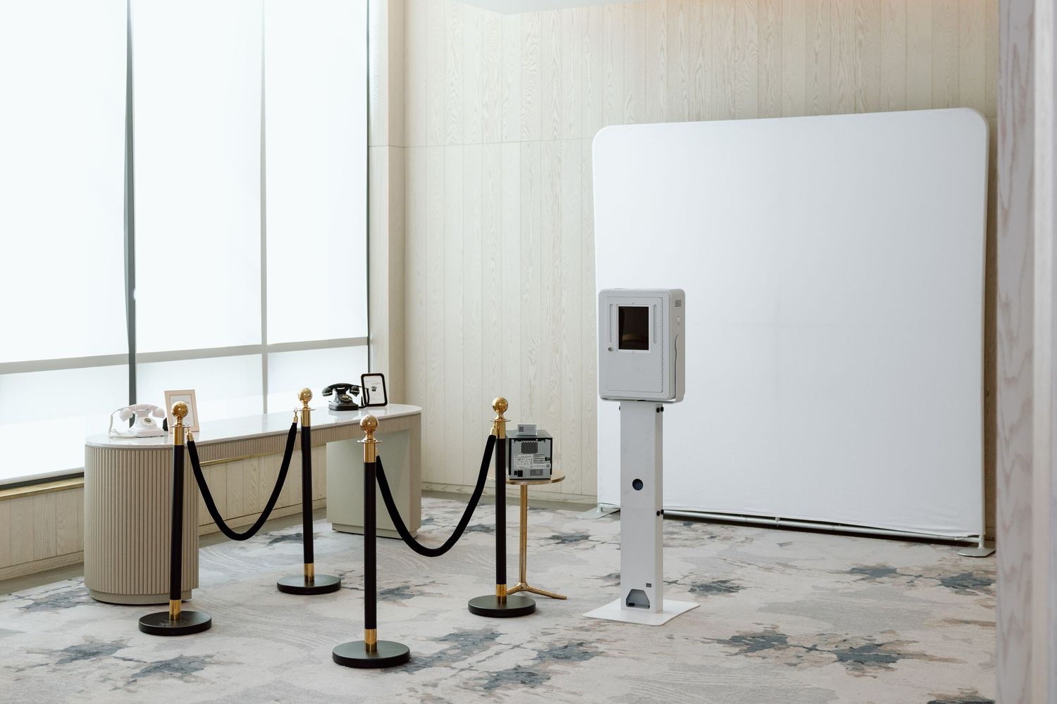 Elegant photo booth setup with white backdrop, velvet rope barriers, and gold stanchions in a modern event venue.