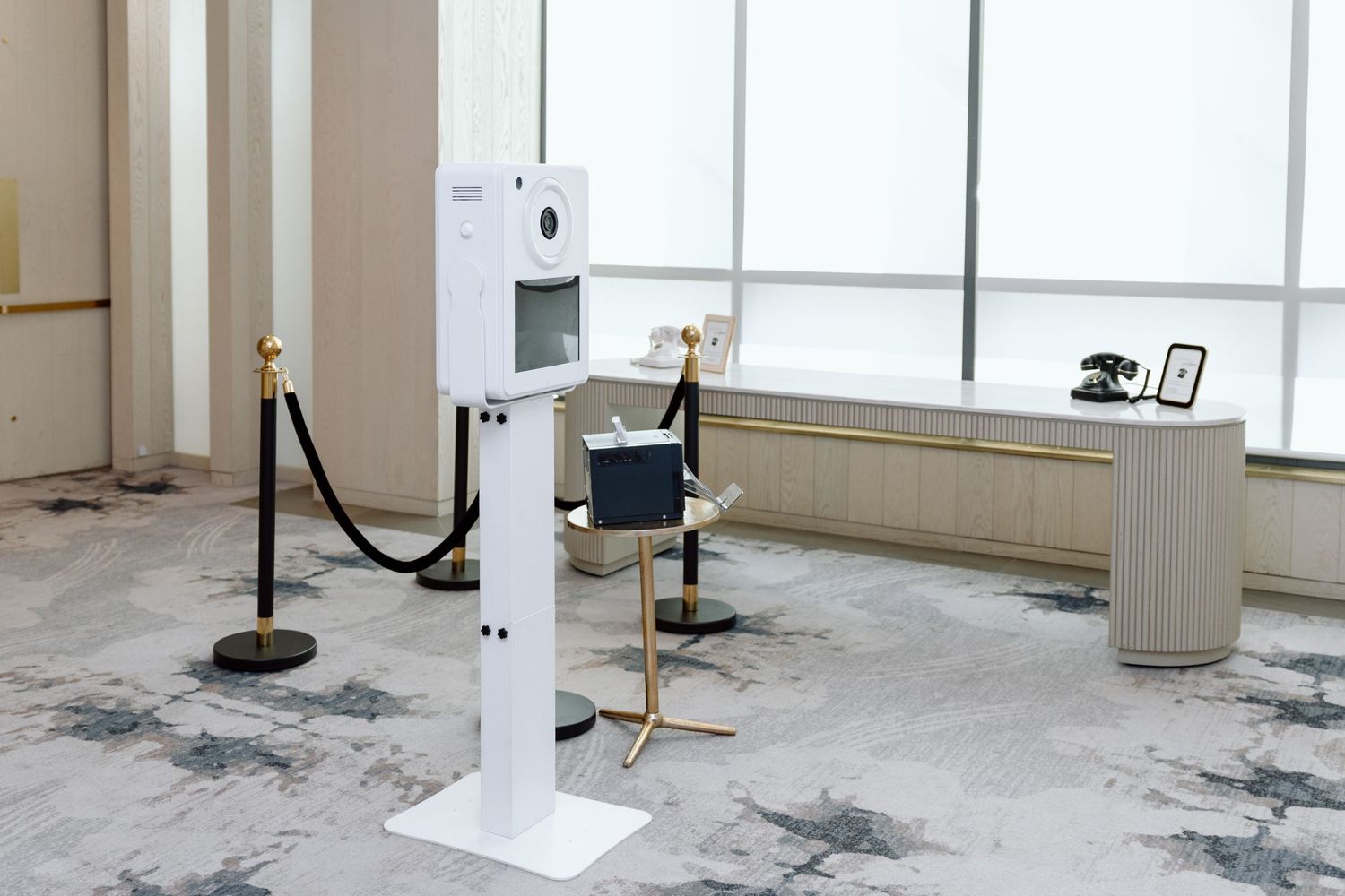 Modern photo booth kiosk on white stand with velvet rope barriers in an elegant event venue.
