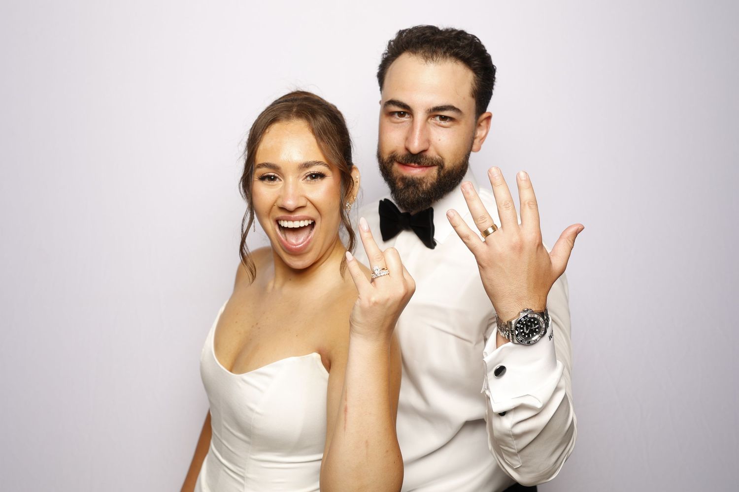 Newlywed couple showing off wedding rings, bride in white gown and groom in tuxedo, smiling at camera.