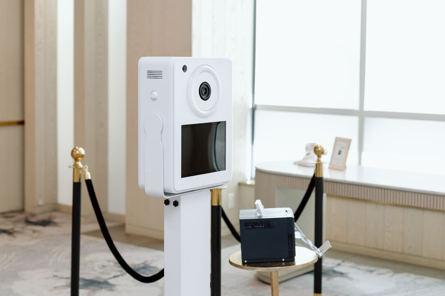 White photo booth kiosk on a pedestal with gold rope barriers in an elegant event venue.