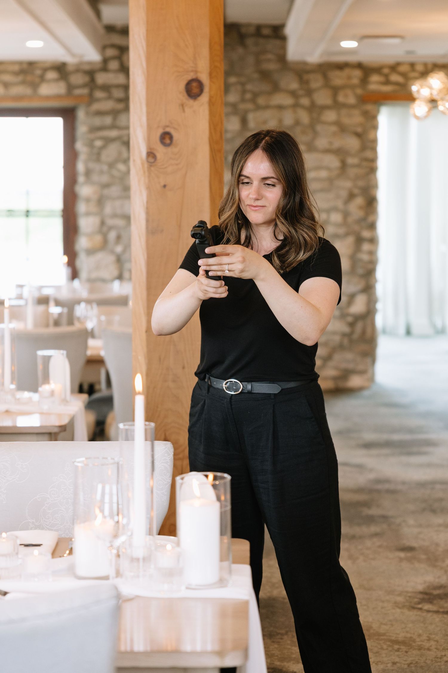 A person in a black outfit arranges white candles and decorative elements on tables in a rustic stone building interior.