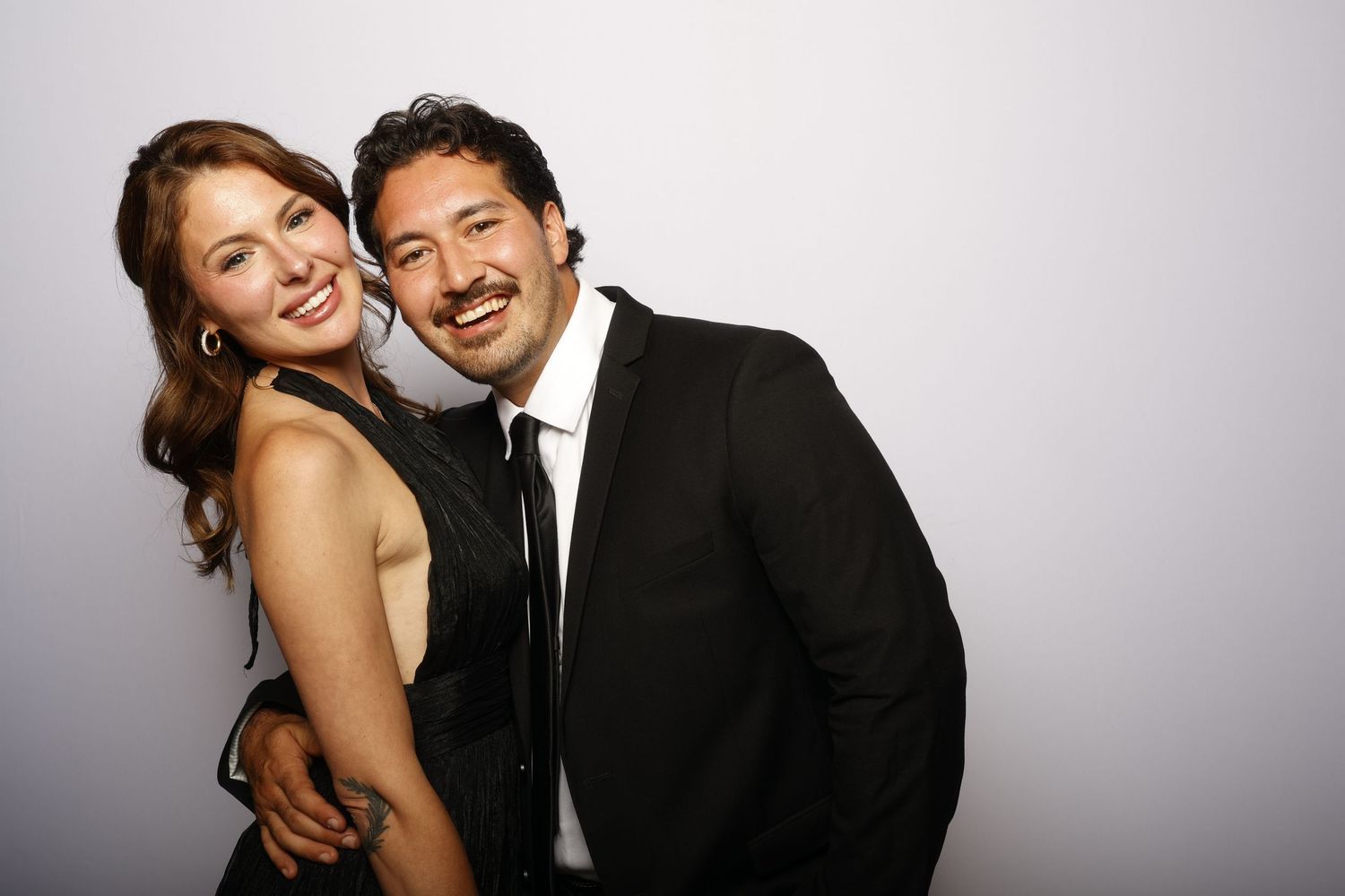 A couple shares a joyful moment together against a gray backdrop, dressed in formal attire.
