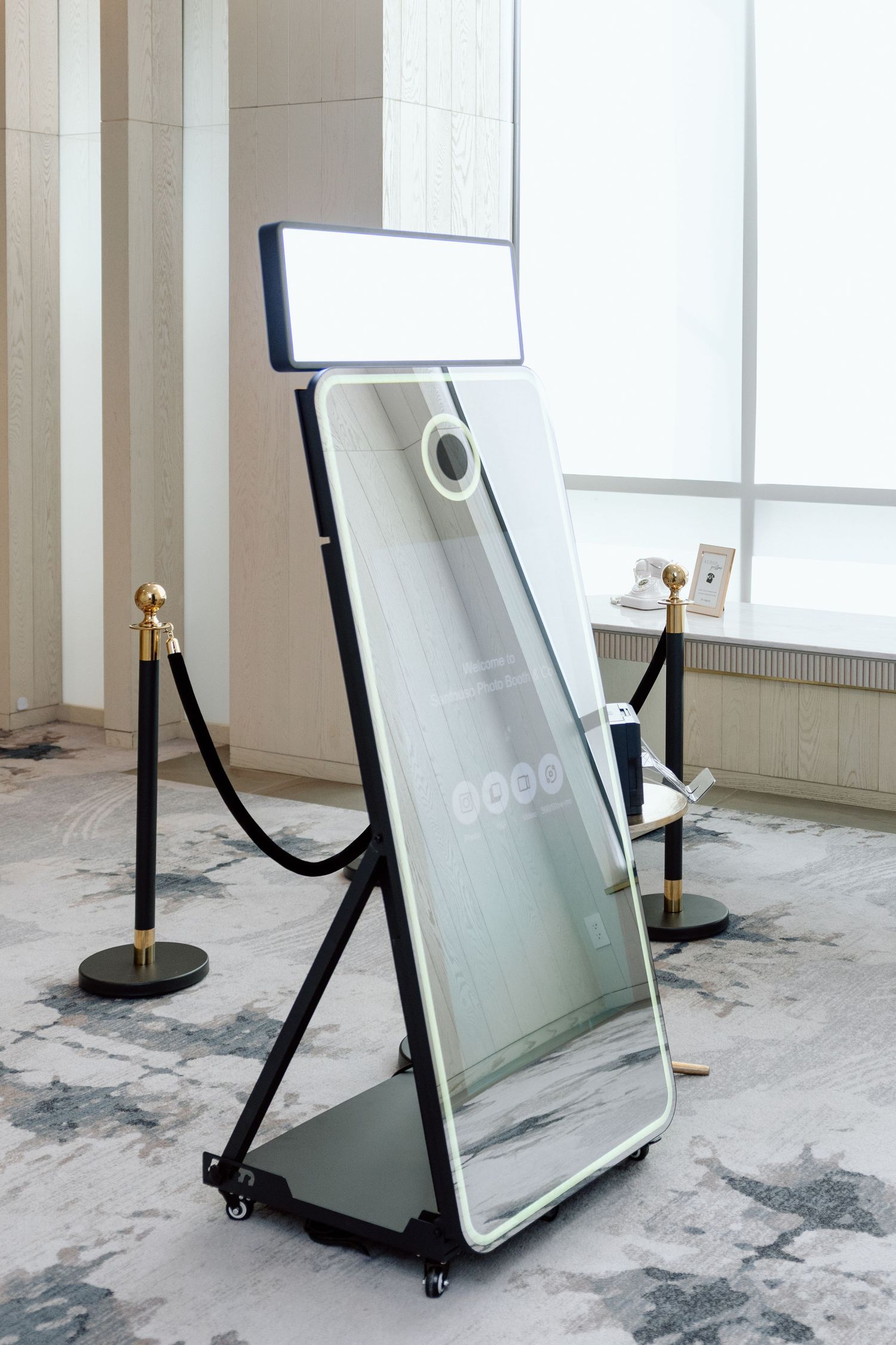 A modern mirror photo booth with LED lighting on a wheeled stand, framed by black velvet rope barriers with gold posts.