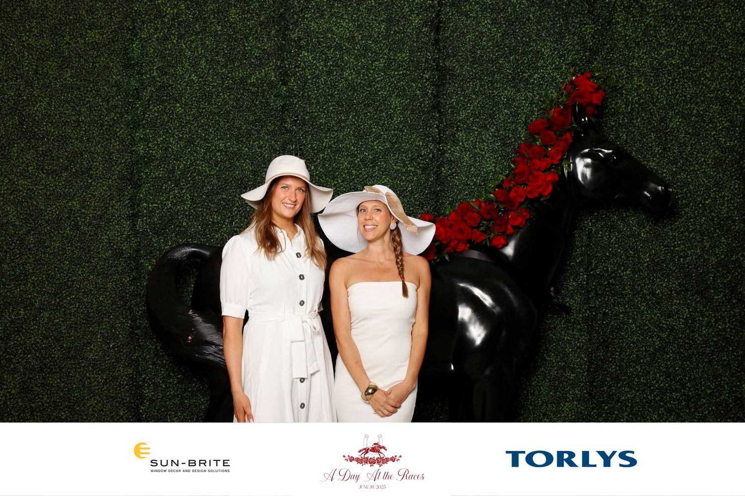 Two women in white sundresses and sun hats pose with a decorative black horse at a Torlys sponsored event.