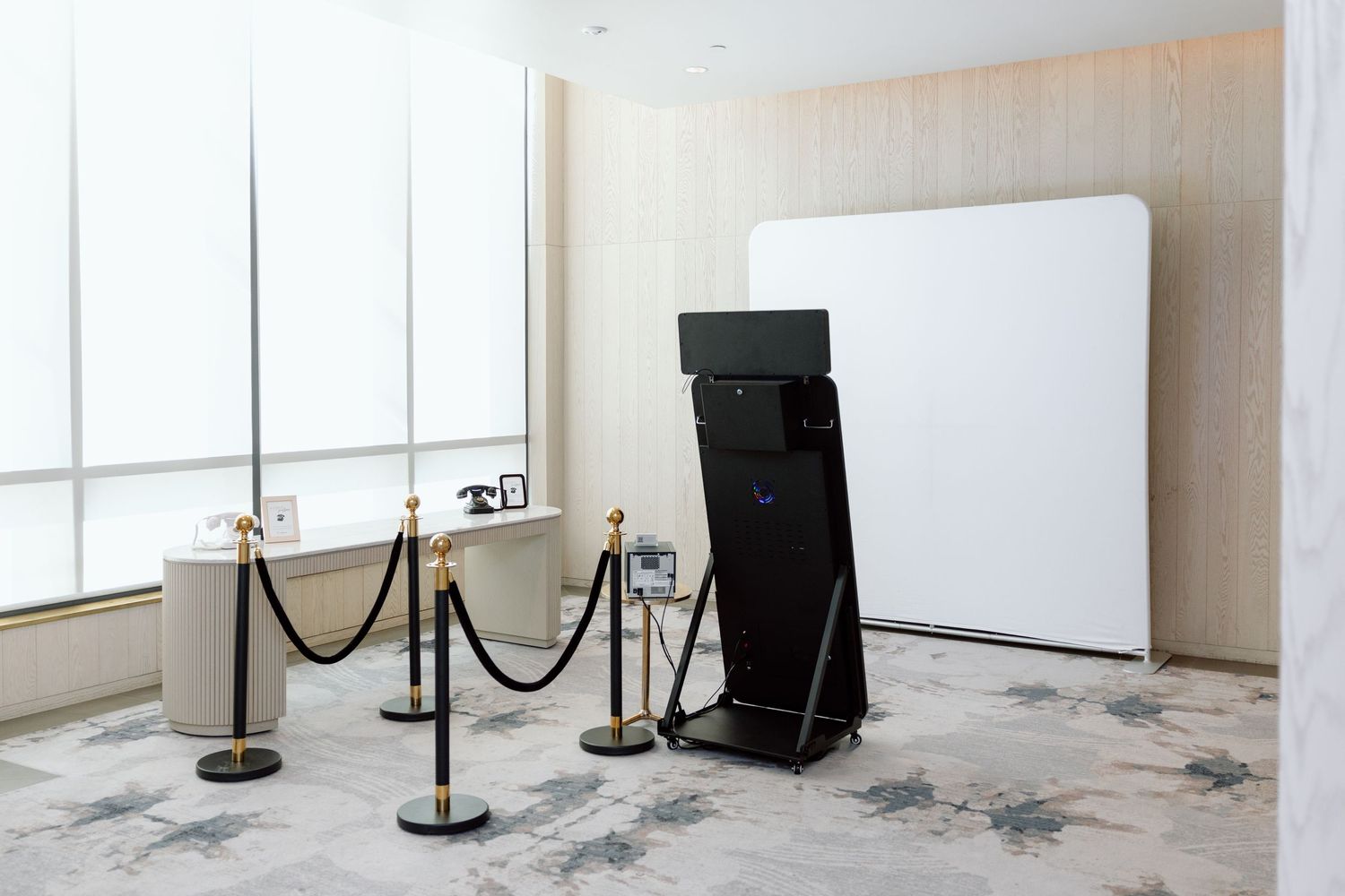 Modern photo booth setup with black kiosk, white backdrop, and gold stanchions in an elegant event space.