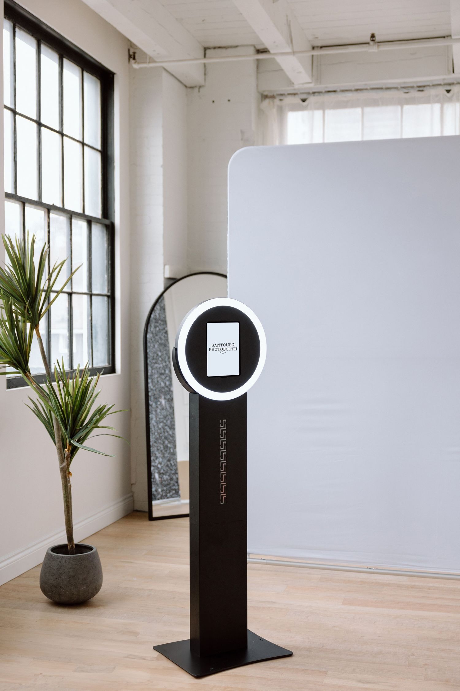 Modern black photo booth kiosk with circular screen display stands next to potted palm plant in bright minimalist room.
